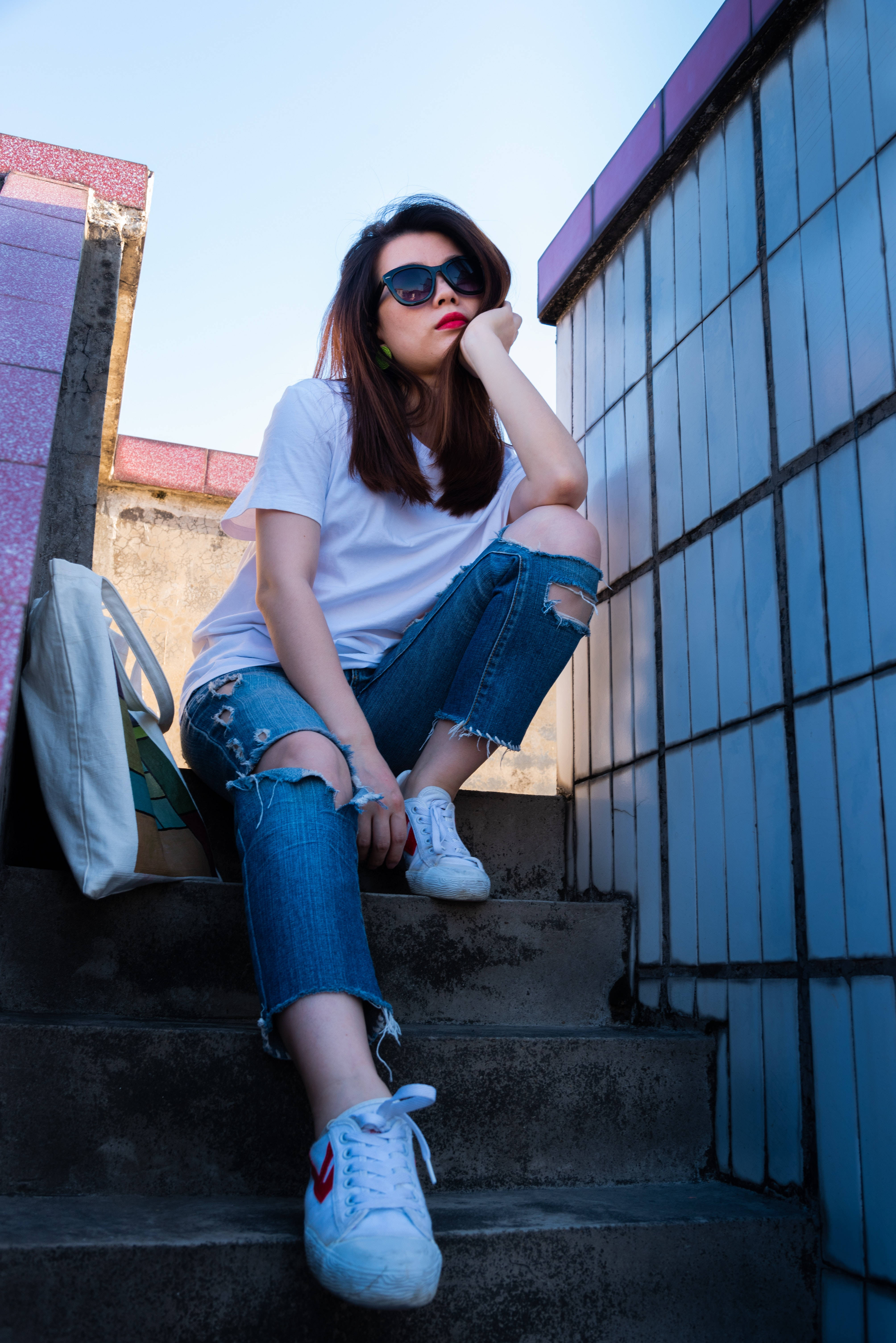女孩 桌布 壁紙 — 一位年輕女性穿著破洞牛仔褲和白色運動鞋，在晴朗的天空下，靠著瓷磚牆，隨意地坐在混凝土台階上。