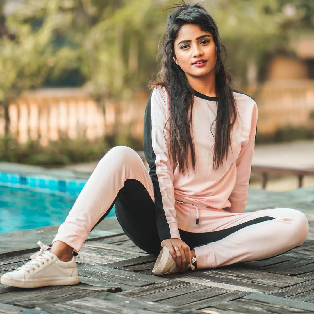 Nisha 桌布 壁紙 — 一位年輕女性穿著粉紅色和黑色的休閒服，隨意地坐在泳池邊的木製甲板上，身後有柔焦的綠色植物。