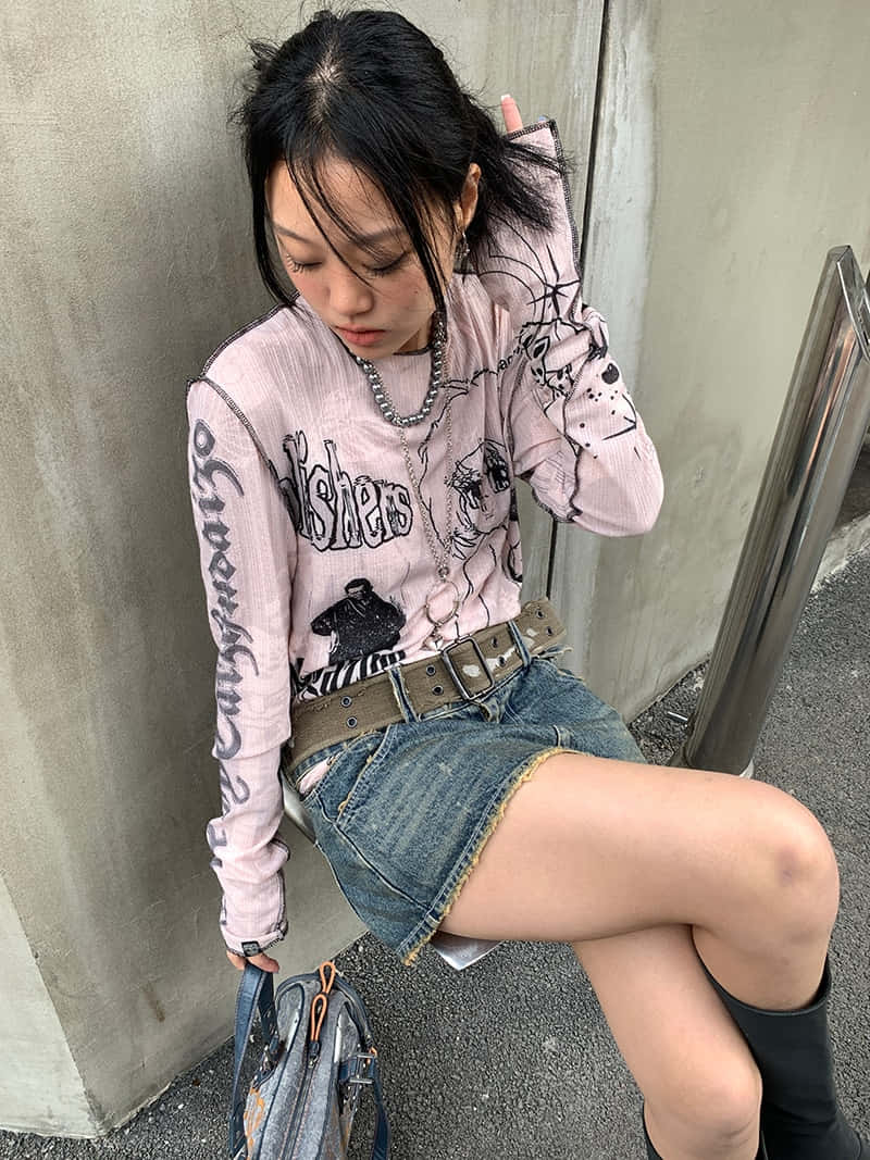 垃圾美學 桌布 壁紙 — 一位年輕女性穿著粉紅色圖案毛衣和牛仔短褲，靠著混凝土牆，手持一個袋子。