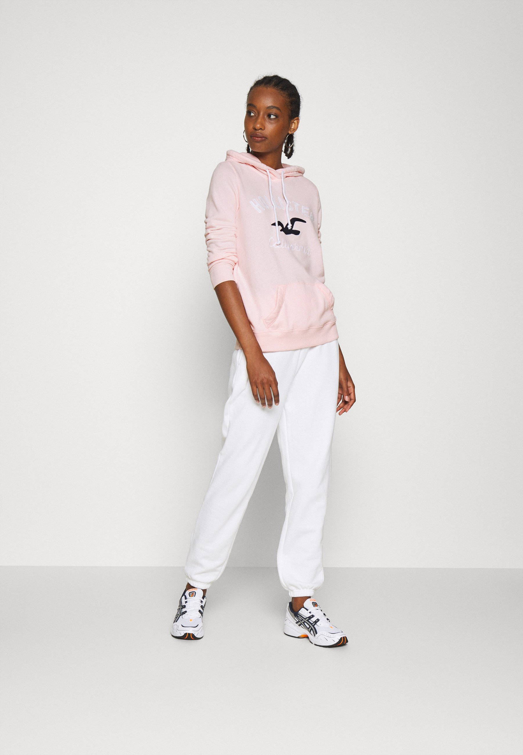 Hollister 桌布 壁紙 — 一位年輕女性穿著粉紅色連帽衫和白色運動褲，自信地站在純白色工作室背景前。