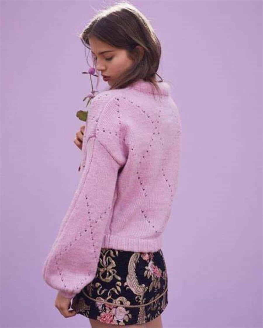 Pink Sweater 桌布 壁紙 — 一位年輕女性穿著粉紅色針織毛衣和花卉裙子，在柔和的薰衣草背景前手持一朵紫色花朵。