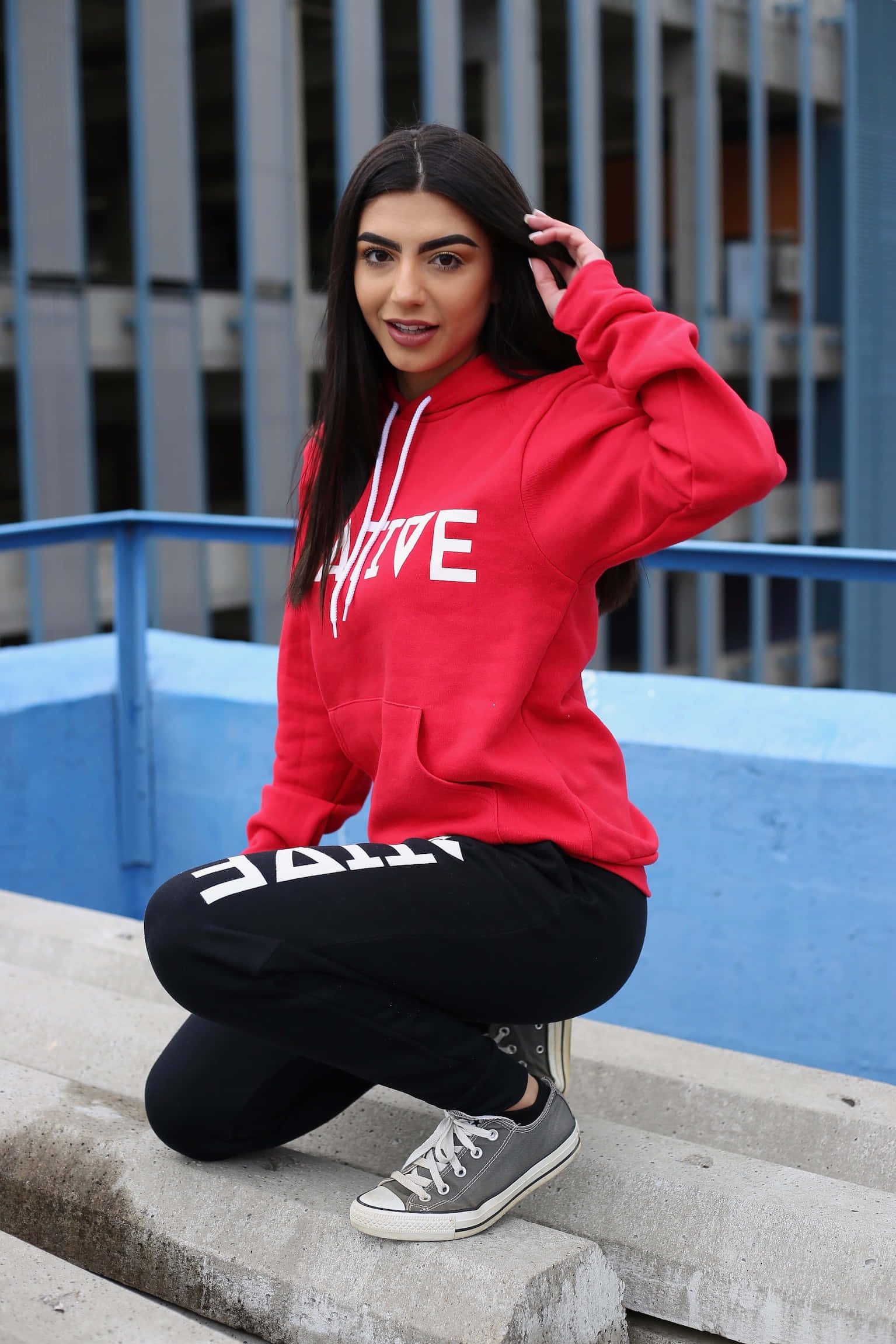 Red Hoodie 桌布 壁紙 — 一位年輕女性穿著紅色連帽衫和黑色慢跑褲，在藍色欄杆和現代建築前的混凝土台階上自信地擺姿勢。