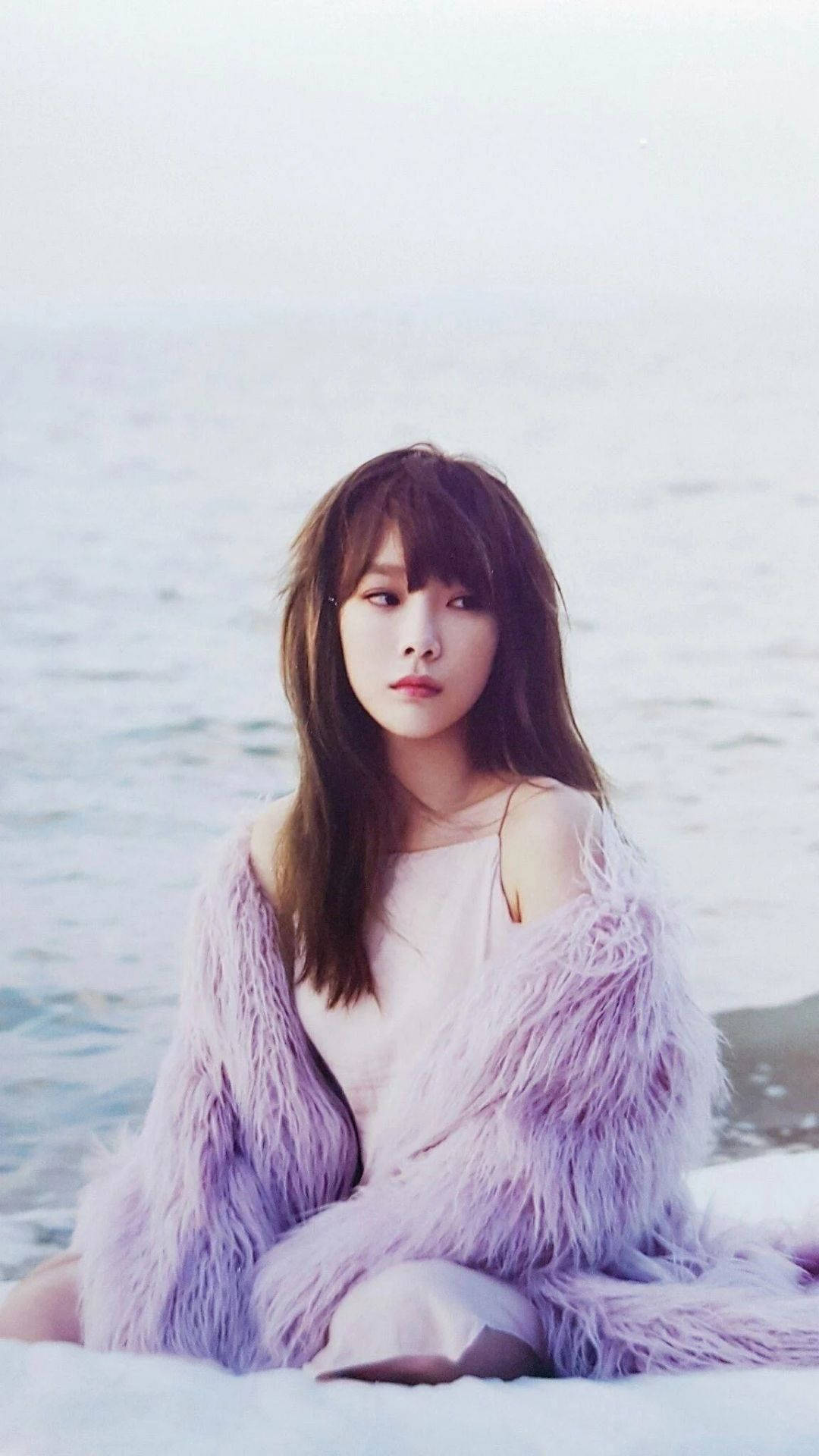 Taeyeon 桌布 壁紙 — 一位年輕女性穿著蓬鬆的薰衣草色外套，在柔和粉彩調和溫柔光線下，平靜地坐在海邊。