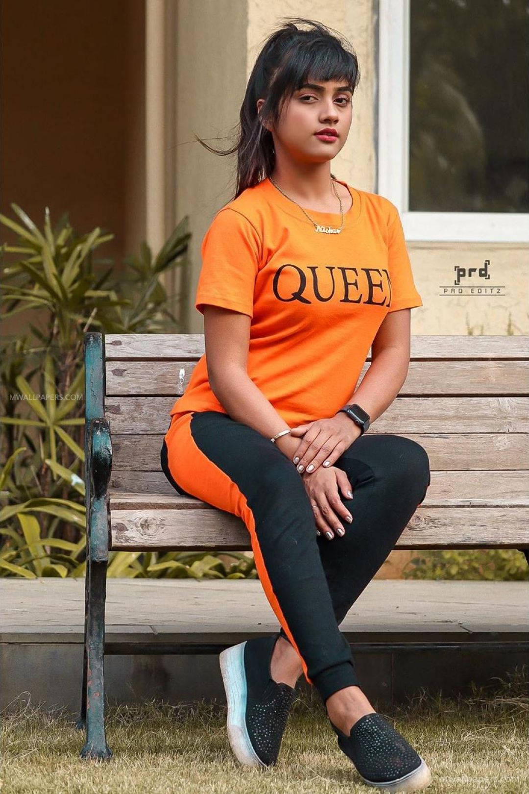 Nisha 桌布 壁紙 — 一位年輕女性穿著鮮橙色 'QUEEN' T恤，自信地坐在戶外一張風化的木長椅上。