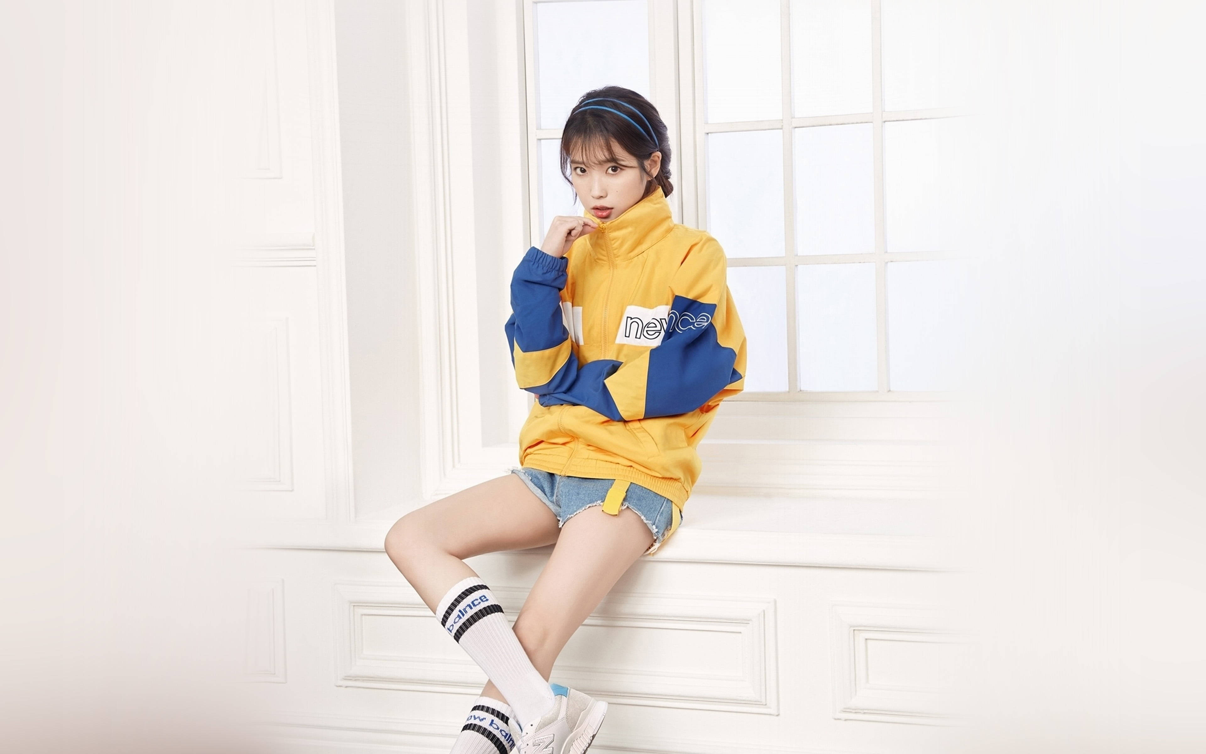 New Balance 桌布 壁紙 — 一位年輕女性穿著黃藍色外套，隨意地坐在白色窗台上，身穿牛仔短褲和條紋襪子。