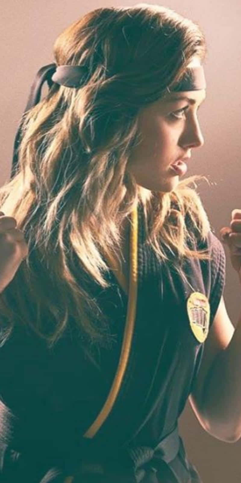 眼鏡蛇道館 桌布 壁紙 — 一位年輕女性穿著黑色武術道服，繫著黃色腰帶和頭帶，在溫暖的側光照射下擺出戰鬥姿勢。