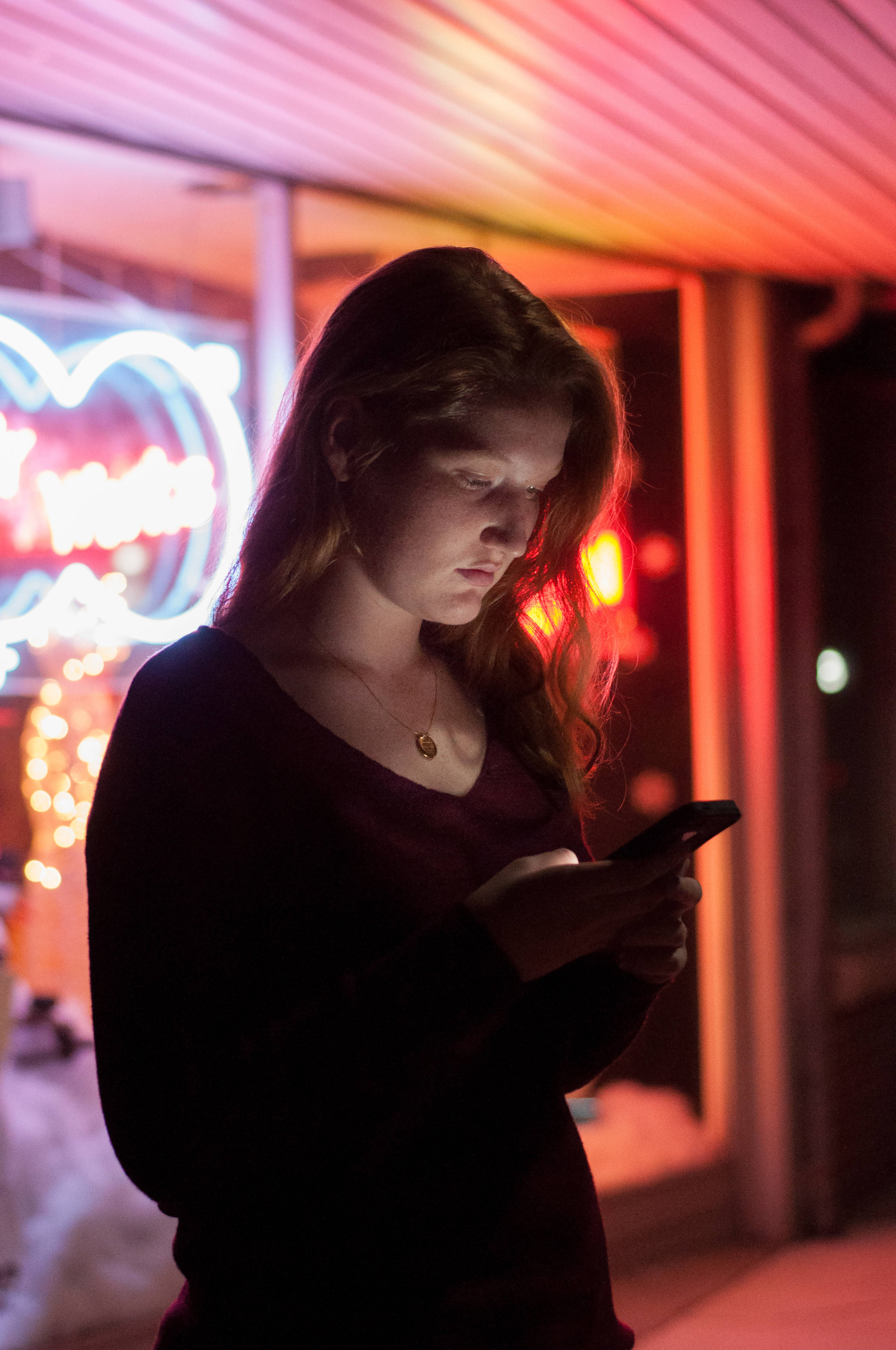 dark neon iPhone 桌布 壁紙 — 一位年輕女性站在霓虹燈照亮的城市夜景中，專注於她的手機，身後色彩繽紛的燈光閃爍。