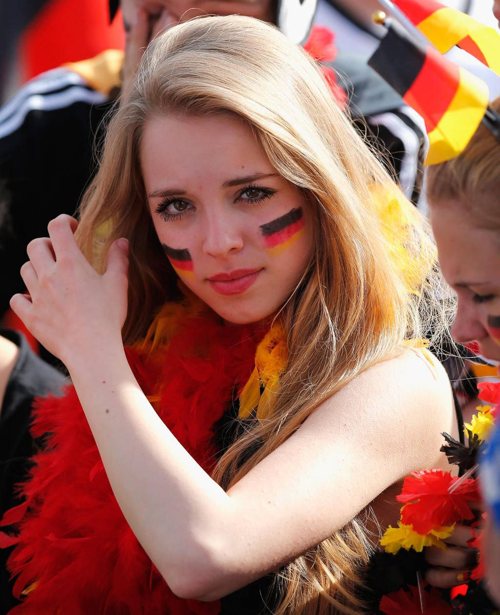 German Girl 桌布 壁紙 — 一位年輕女性臉頰上畫著德國國旗，戴著紅、黃、黑色羽毛圍巾，在熱鬧的戶外活動中微笑。