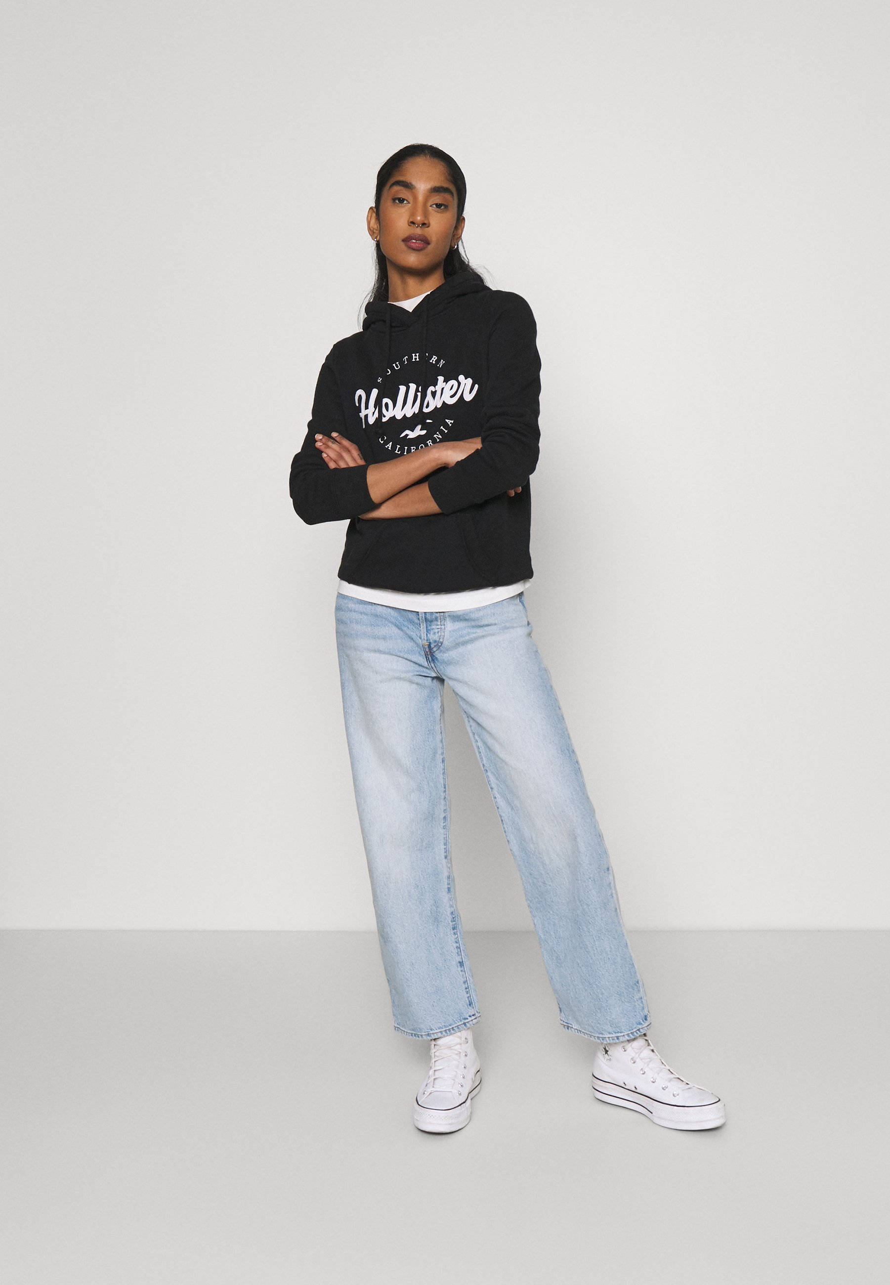 Hollister 桌布 壁紙 — 一位年輕女性自信地交叉雙臂站立，穿著黑色Hollister連帽衫、淺藍色牛仔褲和白色運動鞋，背景是純白色攝影棚。