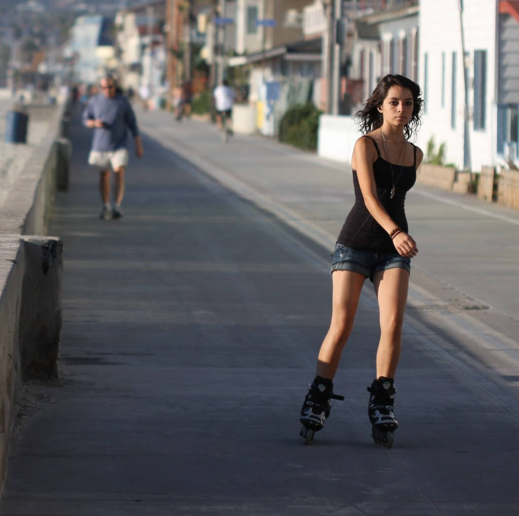 skating 桌布 壁紙 — 一位年輕女性自信地穿著直排輪，沿著陽光普照的海濱長廊滑行，背景是模糊的行人和建築物。