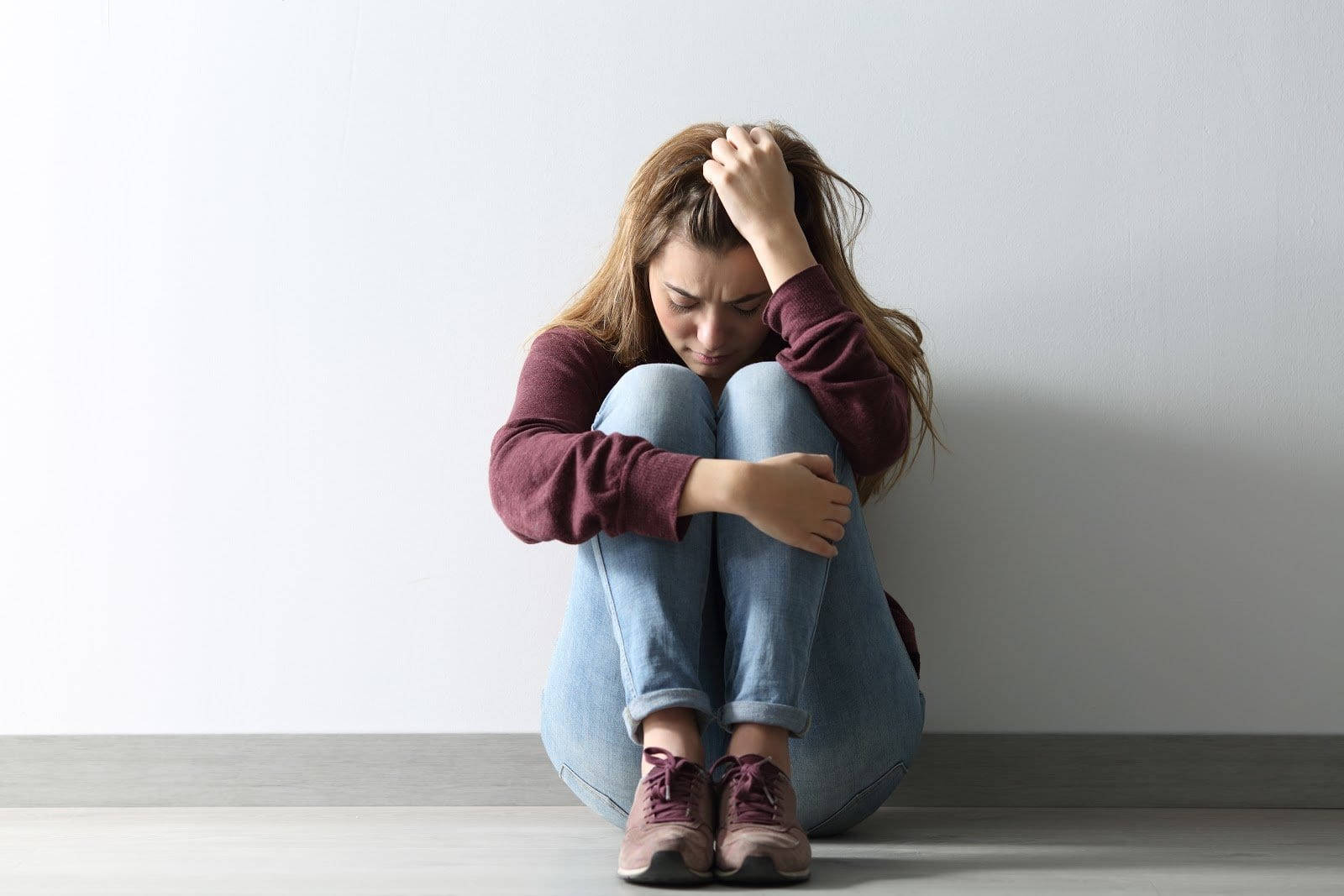 anxious 桌布 壁紙 — 一位年輕女性蜷縮在地上靠著素牆，雙手抱頭，在簡約的室內環境中傳達深深的痛苦。