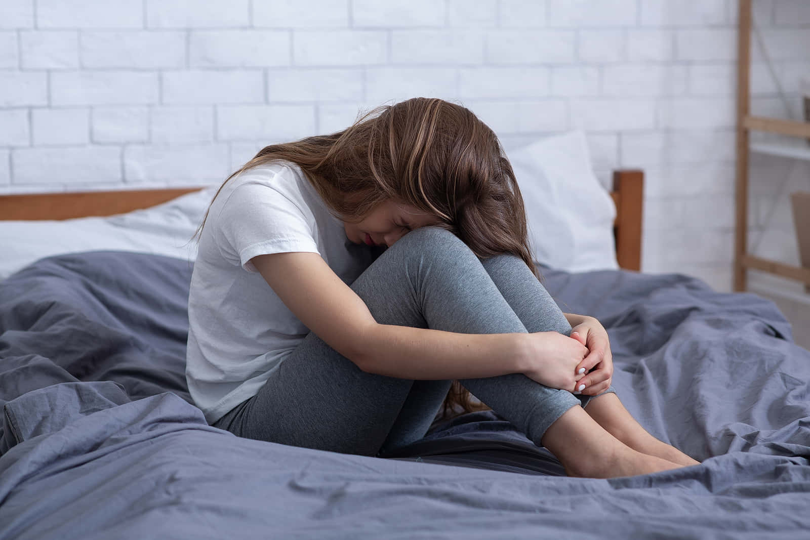 Depressed 桌布 壁紙 — 一位年輕女性蜷縮在床上，低著頭，在簡約風格的臥室中顯得悲傷和退縮。