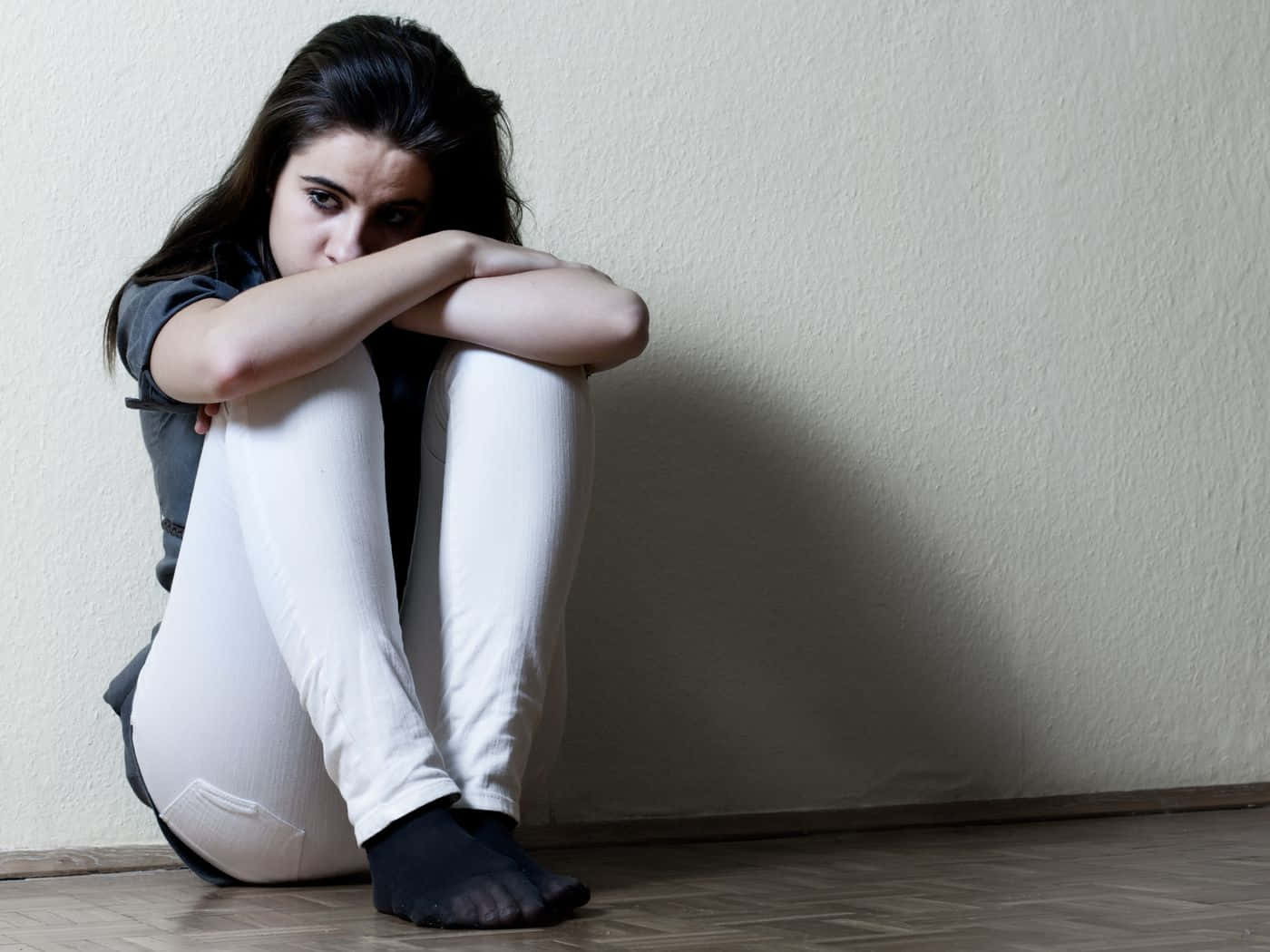 Depressed 桌布 壁紙 — 一位年輕女性蜷縮在木地板上靠著素牆，雙臂環抱膝蓋，眼神流露出深深的悲傷。