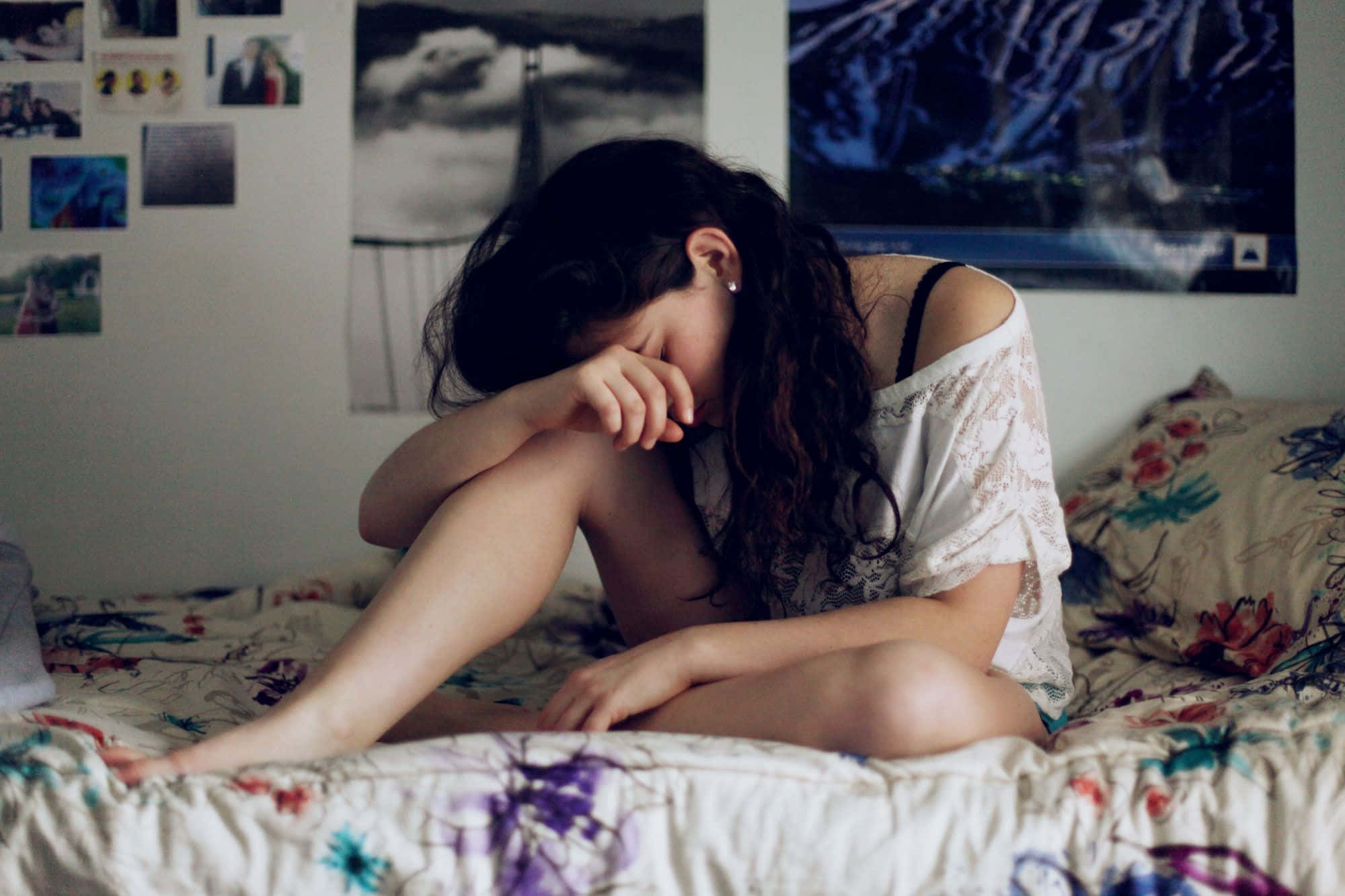 Depression Girl 桌布 壁紙 — 一位年輕女性蜷縮在花卉圖案的床上，雙手抱頭，房間光線昏暗，周圍貼滿牆上海報。
