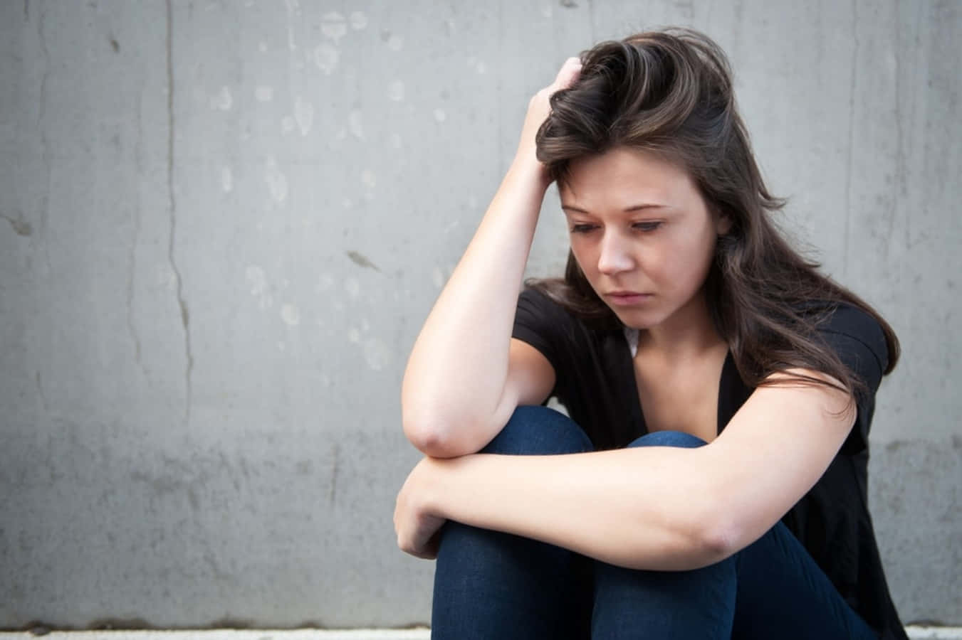 Emotional 桌布 壁紙 — 一位年輕女性蜷縮著坐在灰色混凝土牆邊，手放在頭髮上，低頭帶著悲傷的表情。