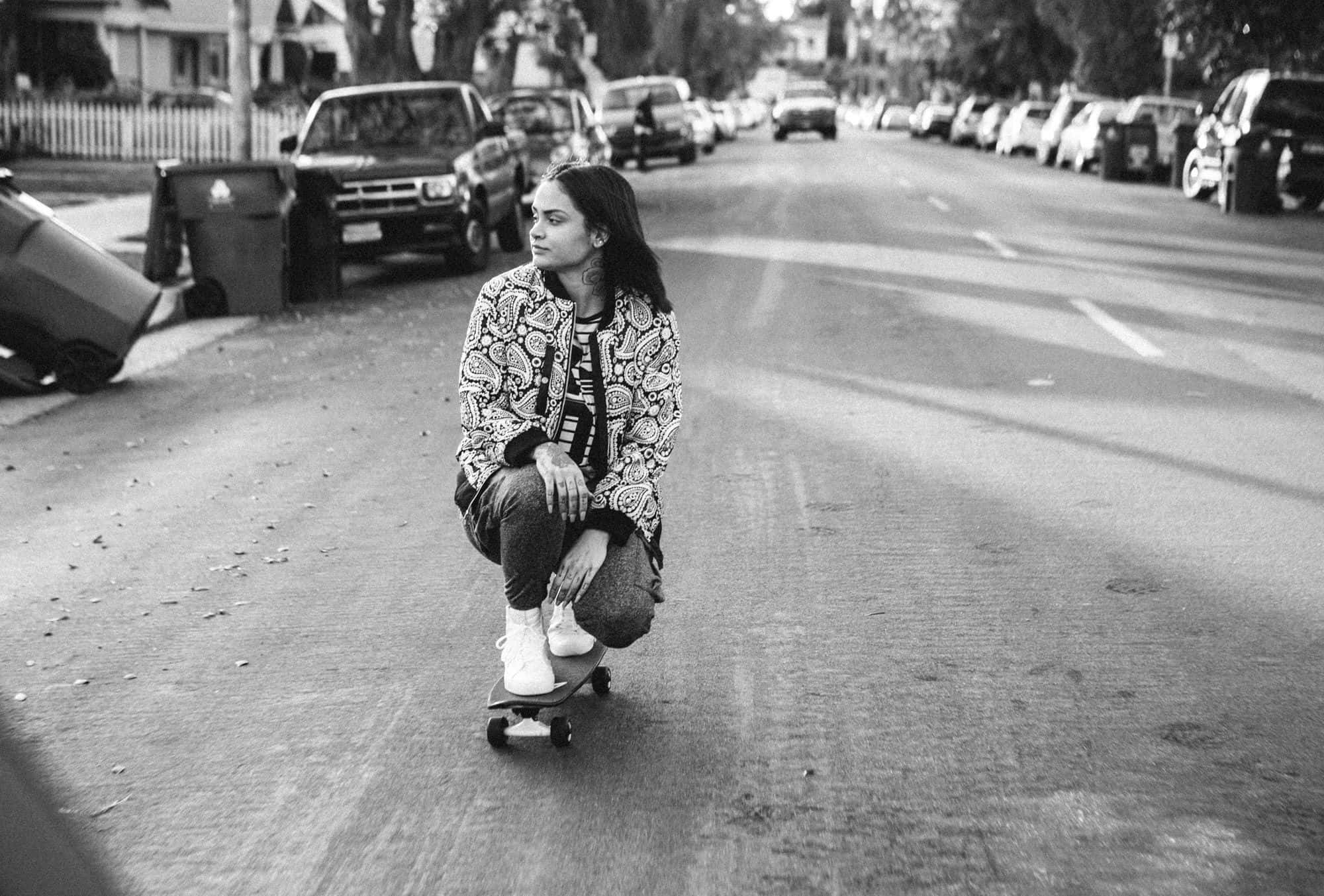 kehlani 桌布 壁紙 — 一位年輕女性蹲在滑板上，攝於寧靜的住宅街道，以黑白呈現。