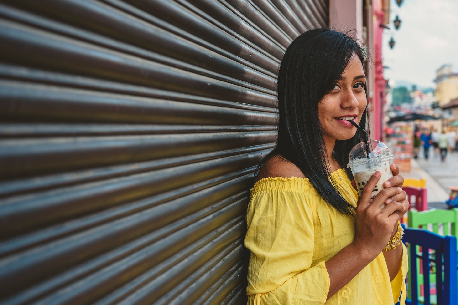 Mexican Woman 桌布 壁紙 — 一位年輕女性身穿亮黃色露肩上衣，在波紋金屬百葉窗前微笑著喝珍珠奶茶。