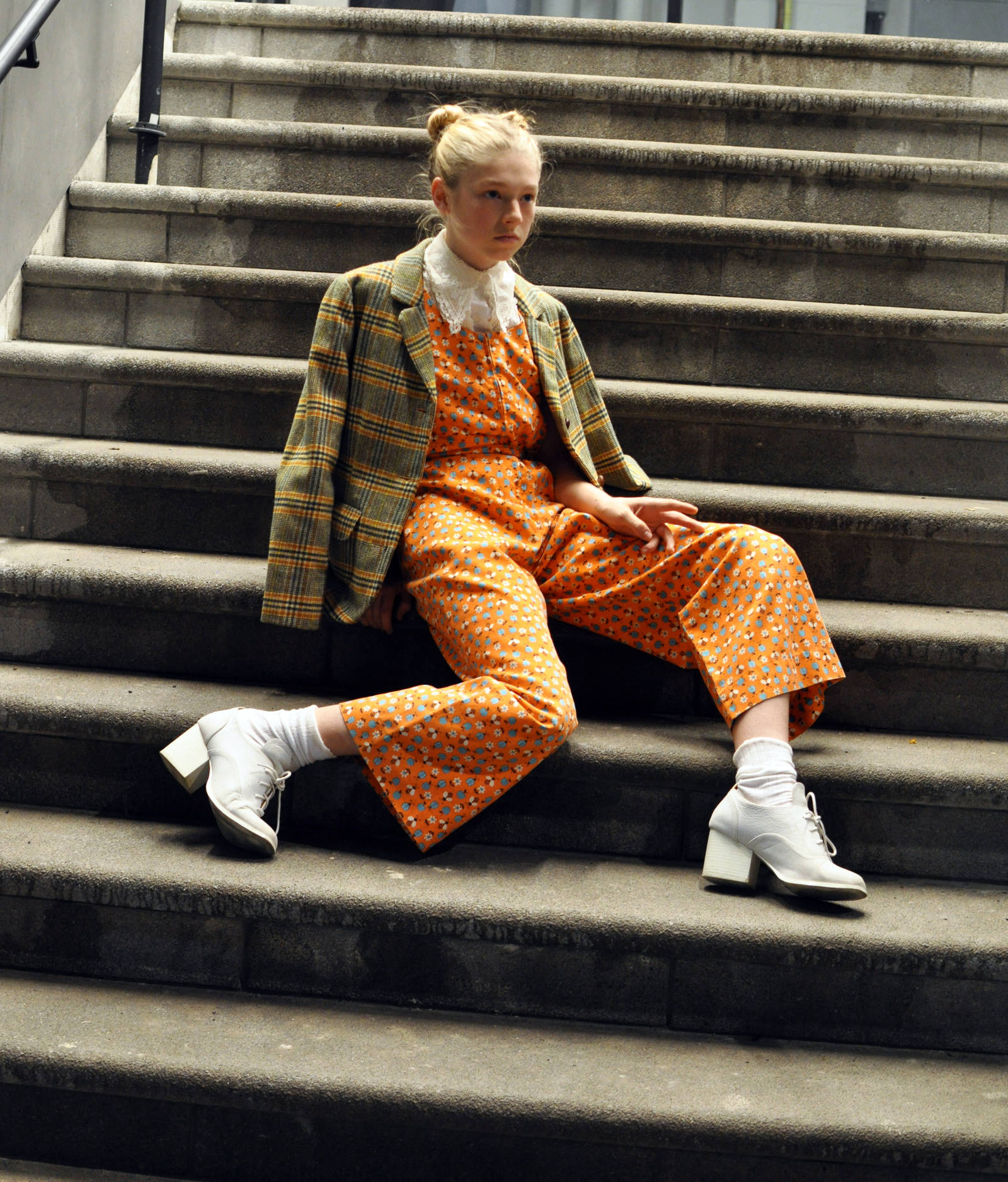 Hunter Schafer 桌布 壁紙 — 一位年輕女性身穿大膽的橙色圖案連身褲和格子西裝外套，從容地坐在風化的石階上。