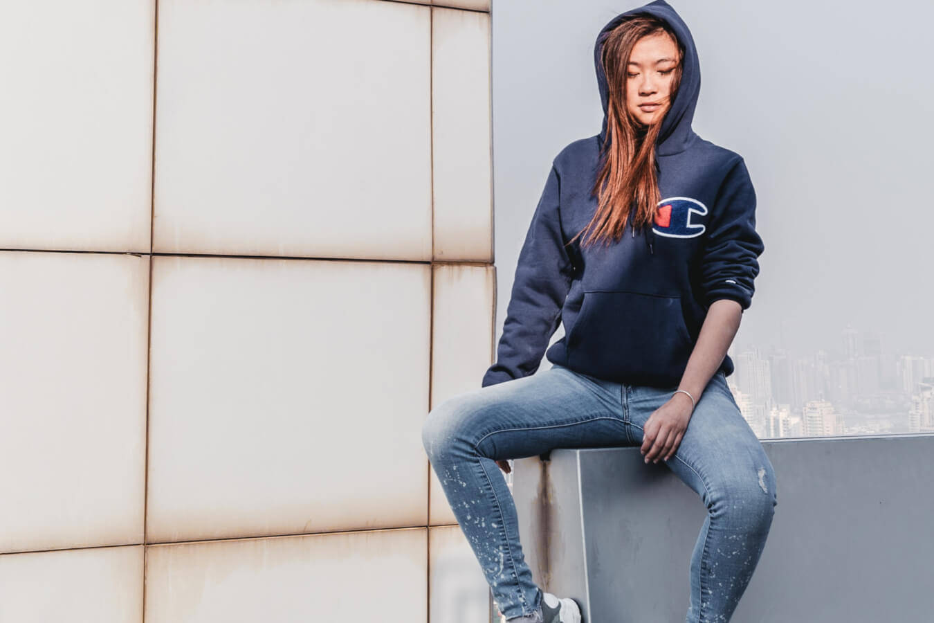 Champion Logo 桌布 壁紙 — 一位年輕女性身穿海軍藍 Champion 連帽衫和破洞牛仔褲，休閒地坐在瓷磚牆旁的屋頂邊緣，身後是朦朧的城市天際線。