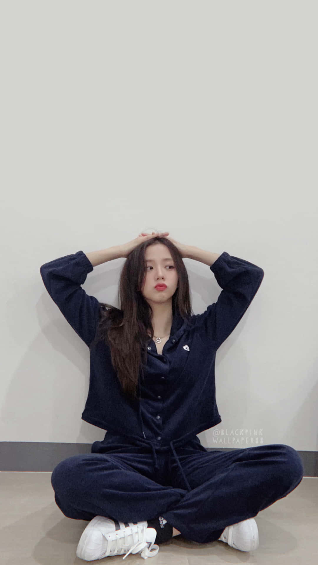 BLACKPINK 桌布 壁紙 — 一位年輕女性身穿海軍藍天鵝絨運動服，雙手放在頭上，盤腿坐在淺色地板上，靠著素牆。