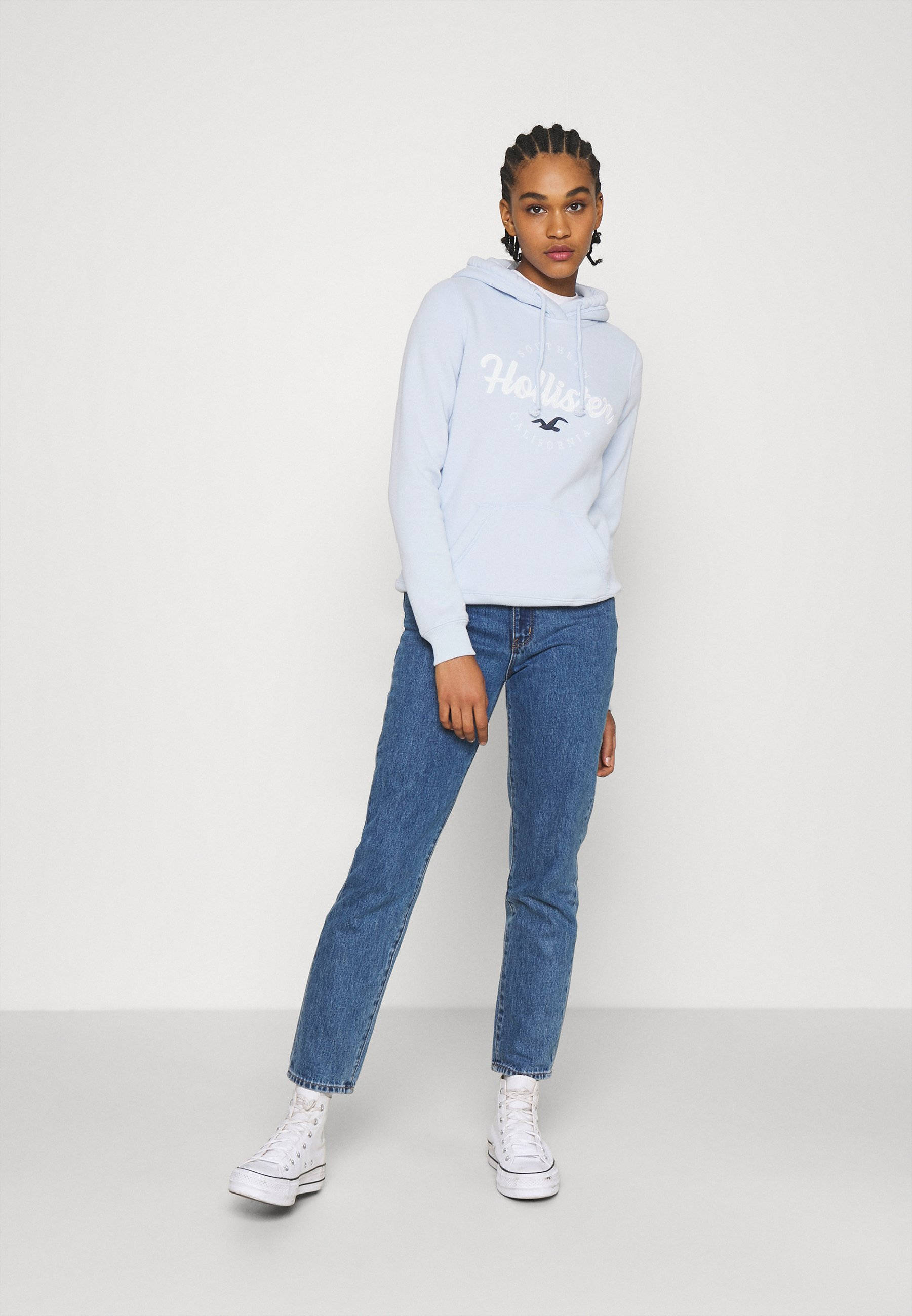 Hollister 桌布 壁紙 — 一位年輕女性身穿淺藍色Hollister連帽衫和藍色牛仔褲，自信地站在純白色攝影棚背景前。