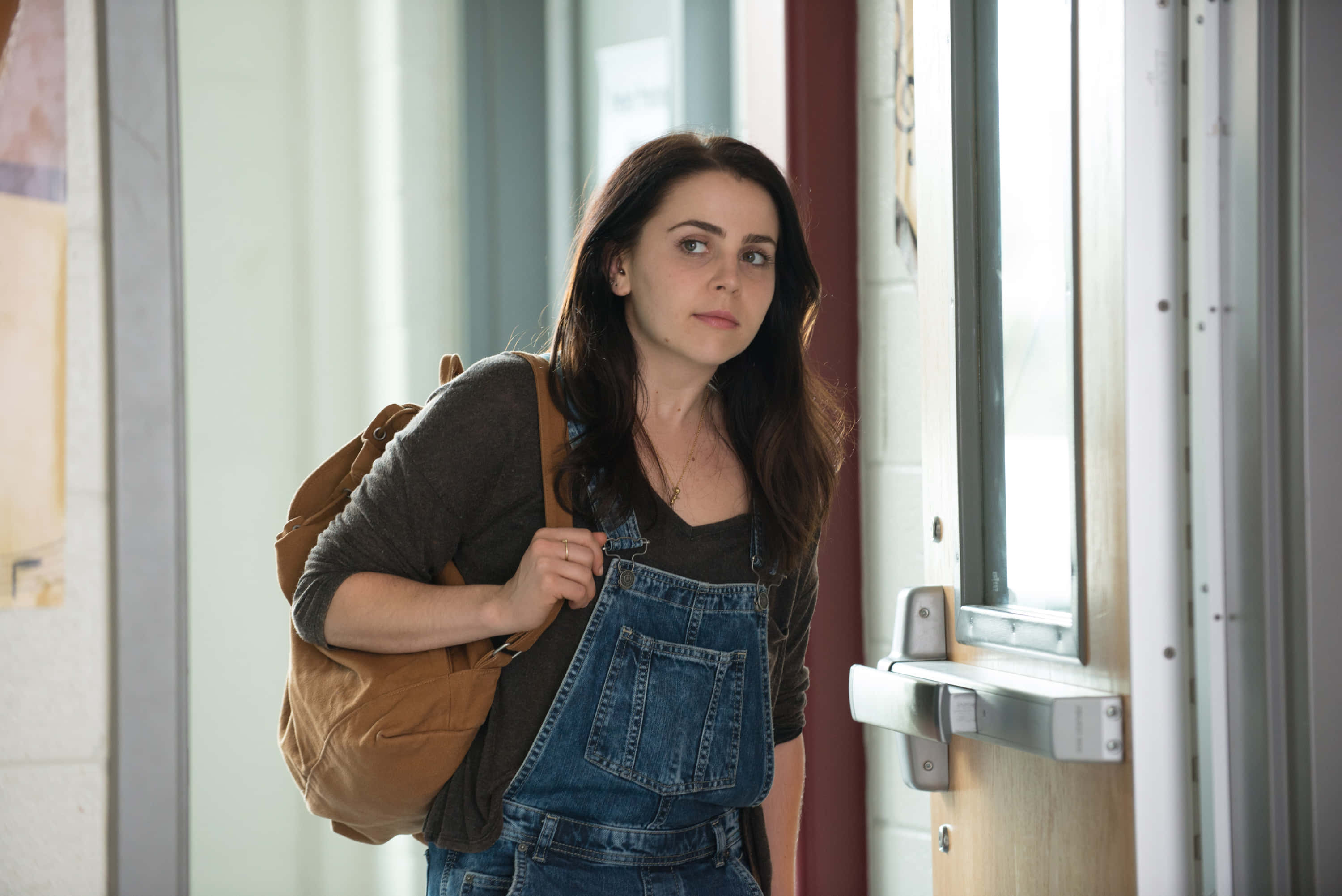 Mae Whitman 桌布 壁紙 — 一位年輕女性身穿牛仔工裝褲和棕色背包，站在學校走廊，若有所思地看向鏡頭。
