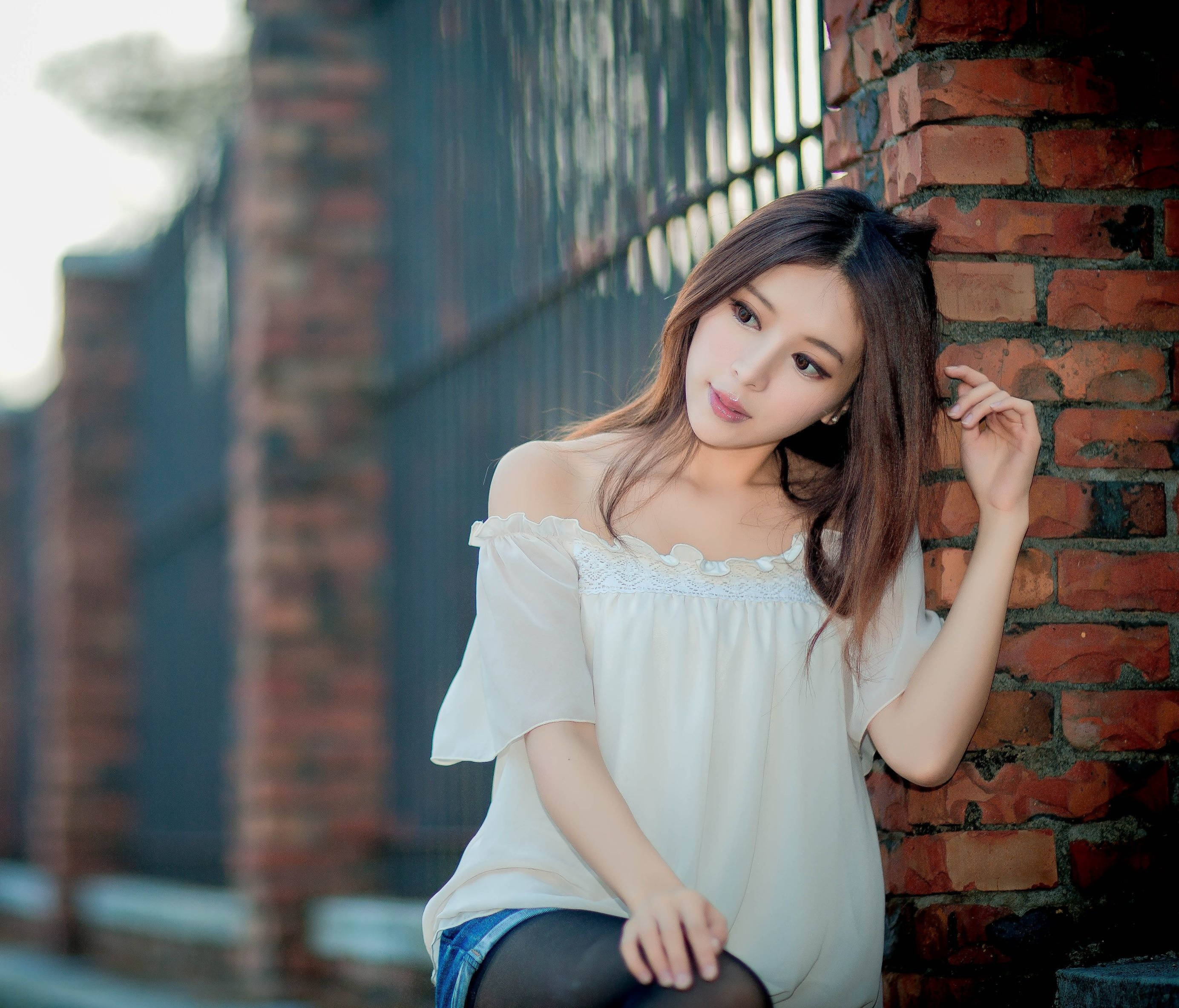 美女 桌布 壁紙 — 一位年輕女性身穿白色露肩衫，靠在風化的磚牆上，柔和自然光照射。
