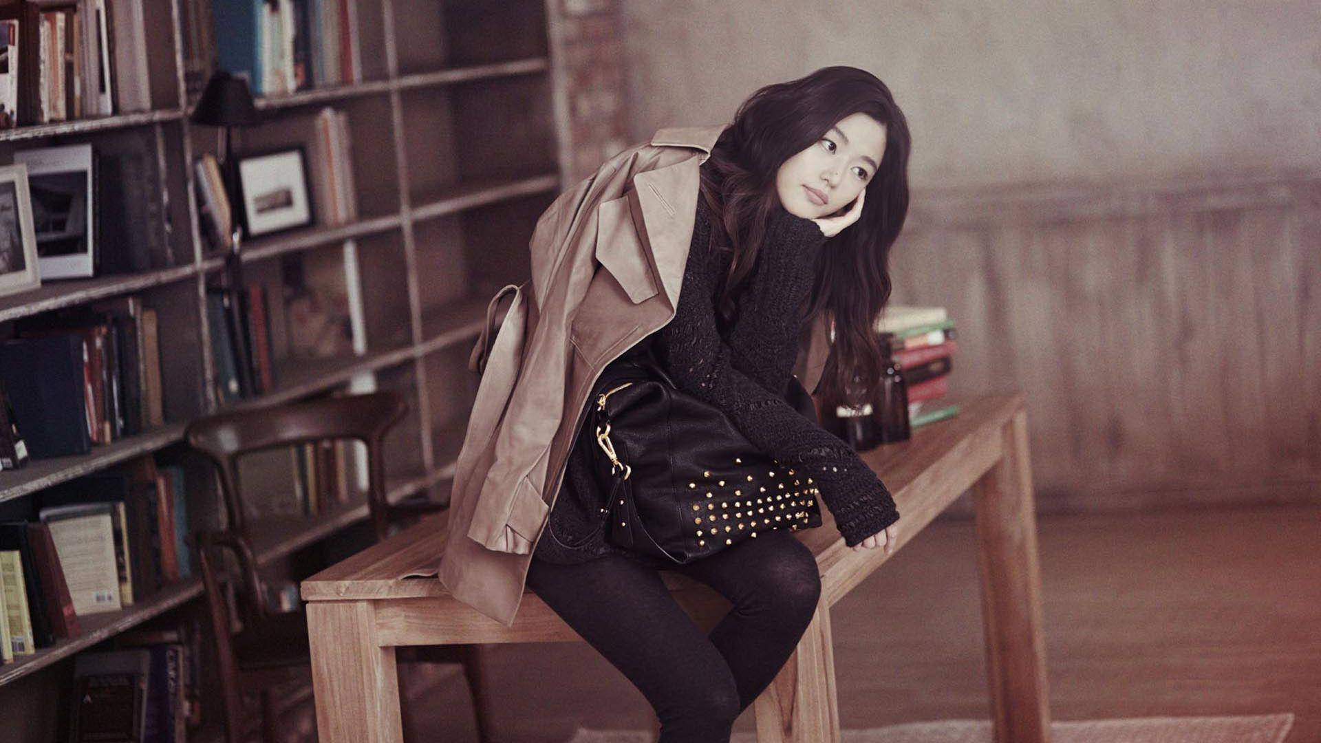 Jun Ji Hyun 桌布 壁紙 — 一位年輕女性身穿米色風衣和黑色針織毛衣，坐在木製長椅上，旁邊是擺滿書籍和裱框照片的書架，若有所思。