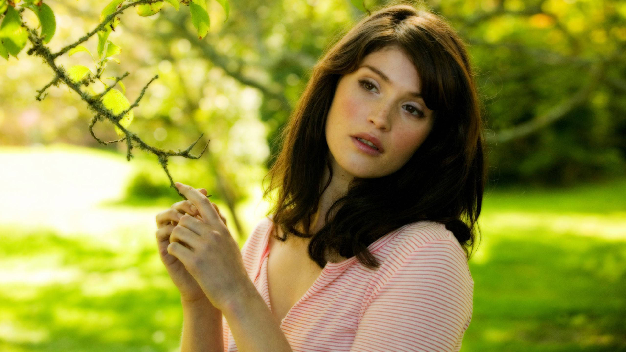 Gemma Arterton 桌布 壁紙 — 一位年輕女性身穿粉紅色條紋襯衫，在陽光普照的綠色花園中輕輕握著一根長滿苔蘚的樹枝。