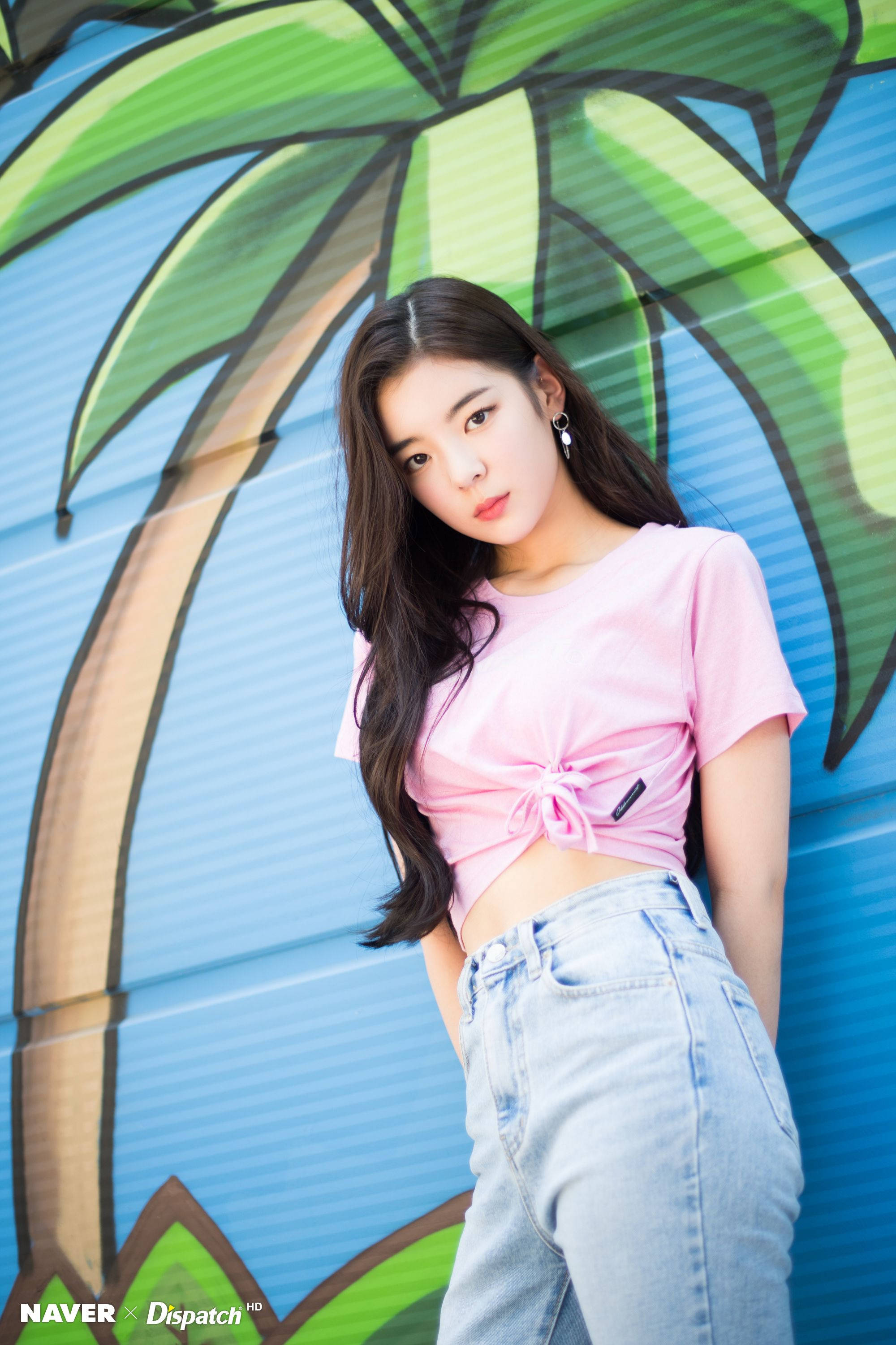 Lia 桌布 壁紙 — 一位年輕女性身穿粉紅色綁帶露臍上衣和淺藍色牛仔褲，在一個帶有大型綠色棕櫚樹壁畫的鮮豔藍色牆壁前自信地擺姿勢。