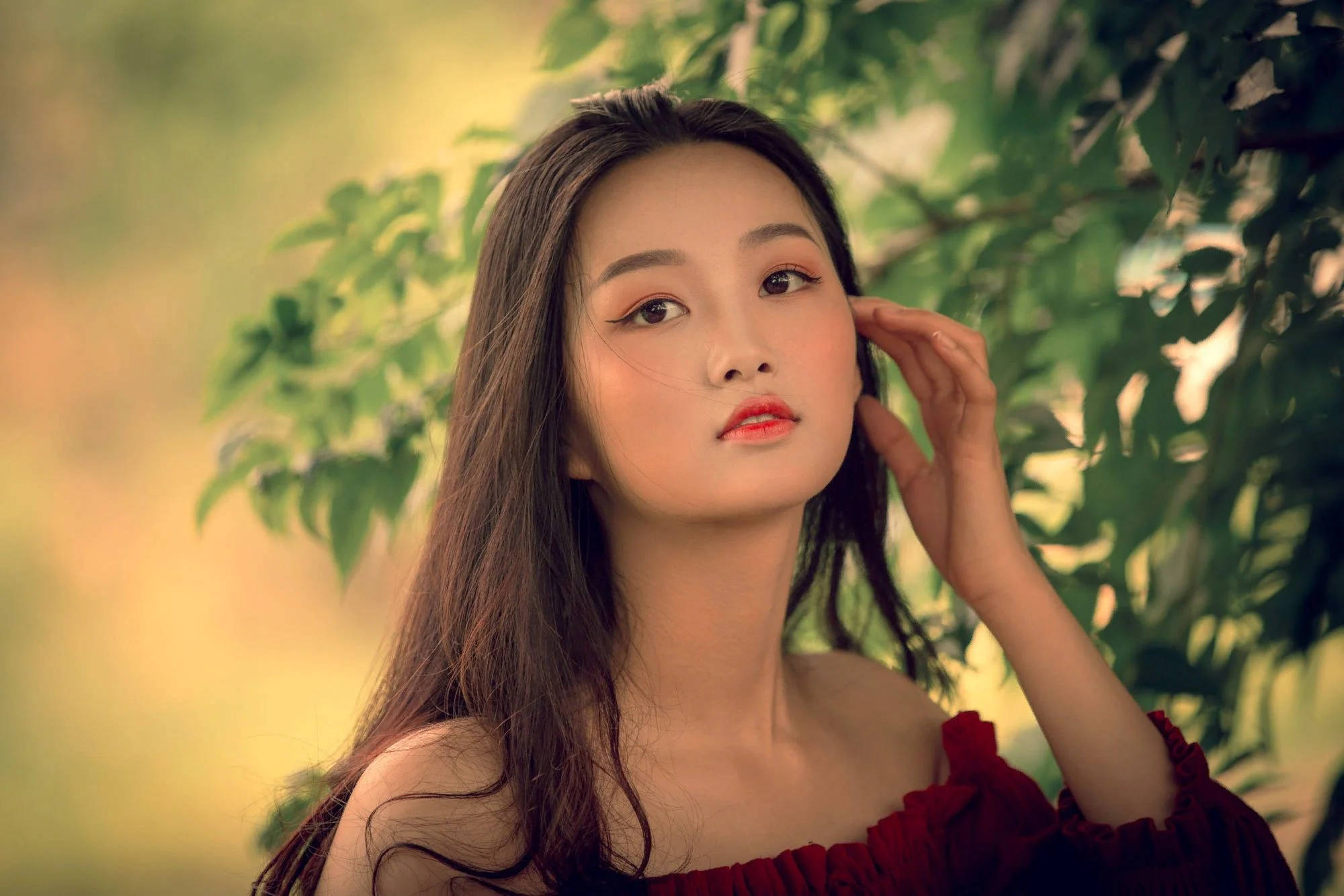 美女 桌布 壁紙 — 一位年輕女性身穿紅色露肩連衣裙，在柔和綠色樹葉下，溫暖金色光線中輕柔觸摸頭髮。