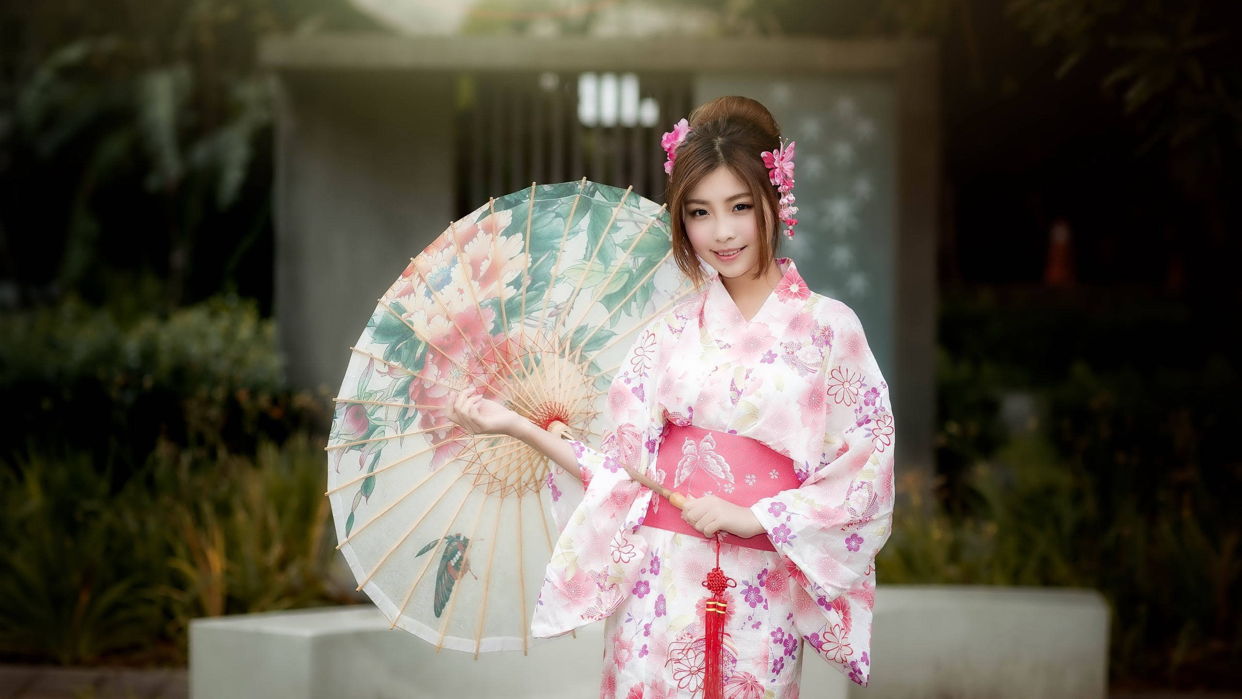 日本 桌布 壁紙 — 一位年輕女性身穿花卉粉色和服，手持相配的紙傘，在茂盛的花園中溫柔微笑。