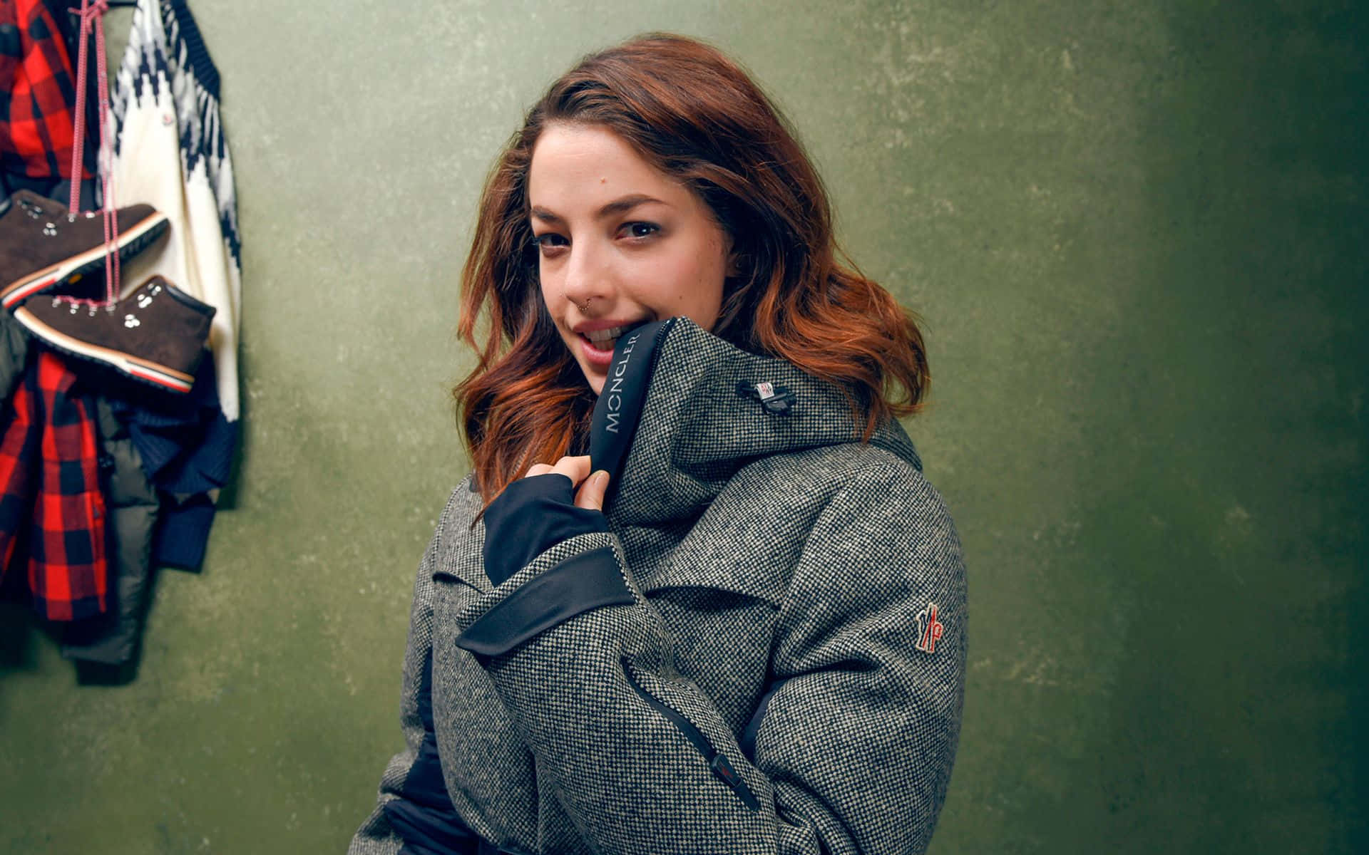 Olivia Thirlby 桌布 壁紙 — 一位年輕女性身穿質感灰色Moncler外套，輕柔微笑，同時將帽子靠向掛有冬季裝備的柔和綠色牆壁。