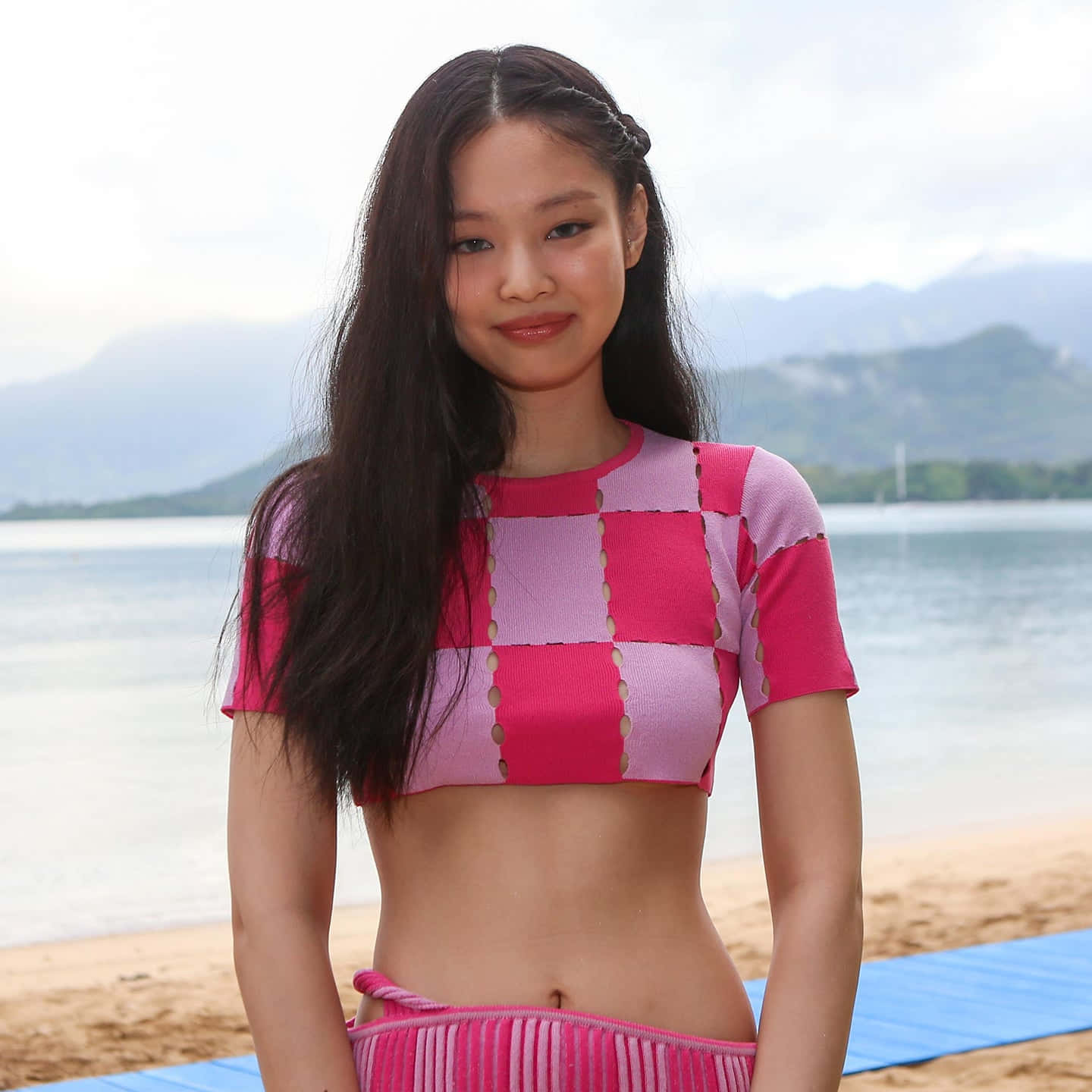 BLACKPINK 桌布 壁紙 — 一位年輕女性身穿鮮豔的粉紅色格紋短版上衣，站在沙灘上，背景是平靜的海水和遠處的山脈，天空多雲。