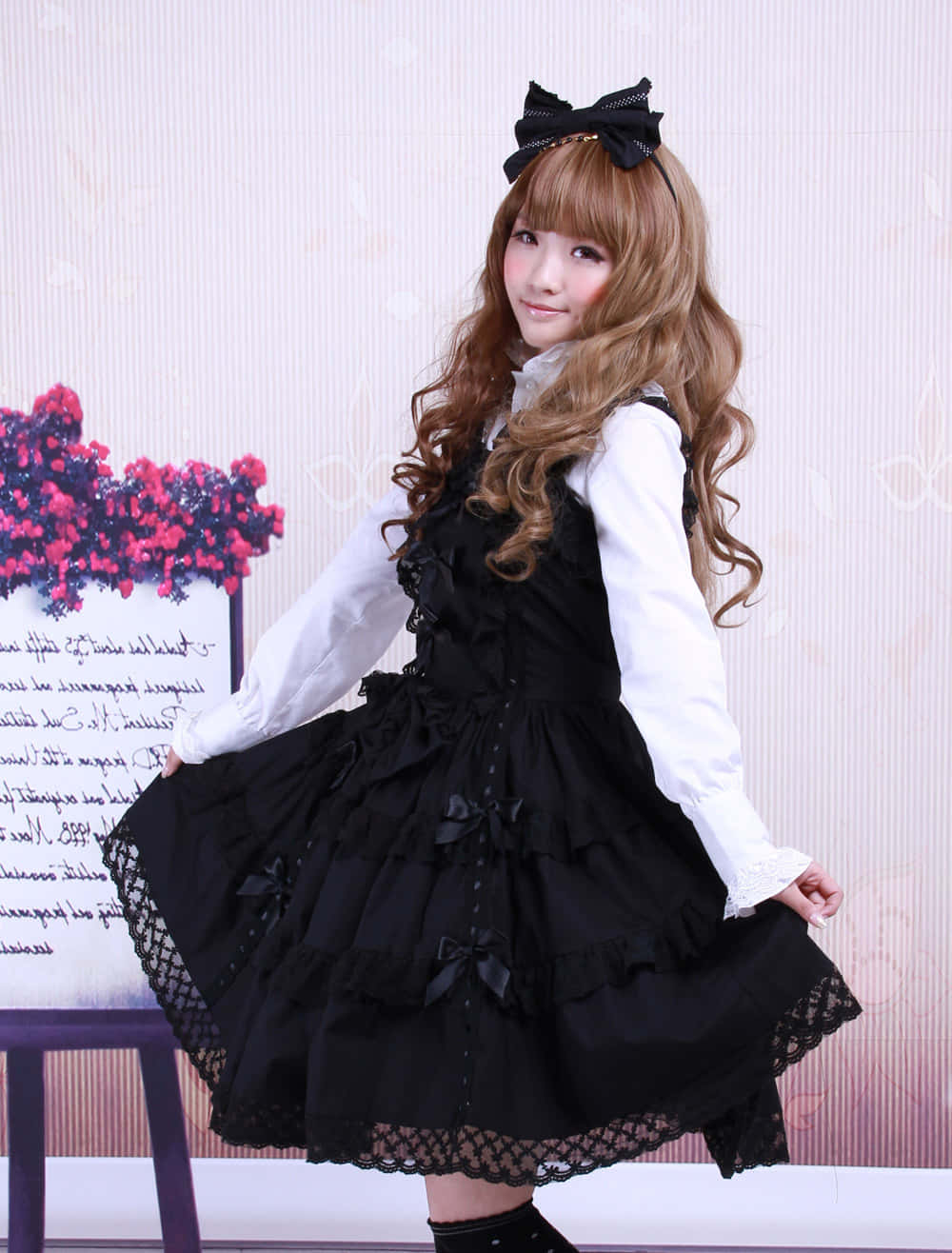 Lolita Fashion 桌布 壁紙 — 一位年輕女性身穿黑色蕾絲Lolita連衣裙，配有蝴蝶結和相配的頭帶，在花卉擺設旁優雅地擺姿勢。