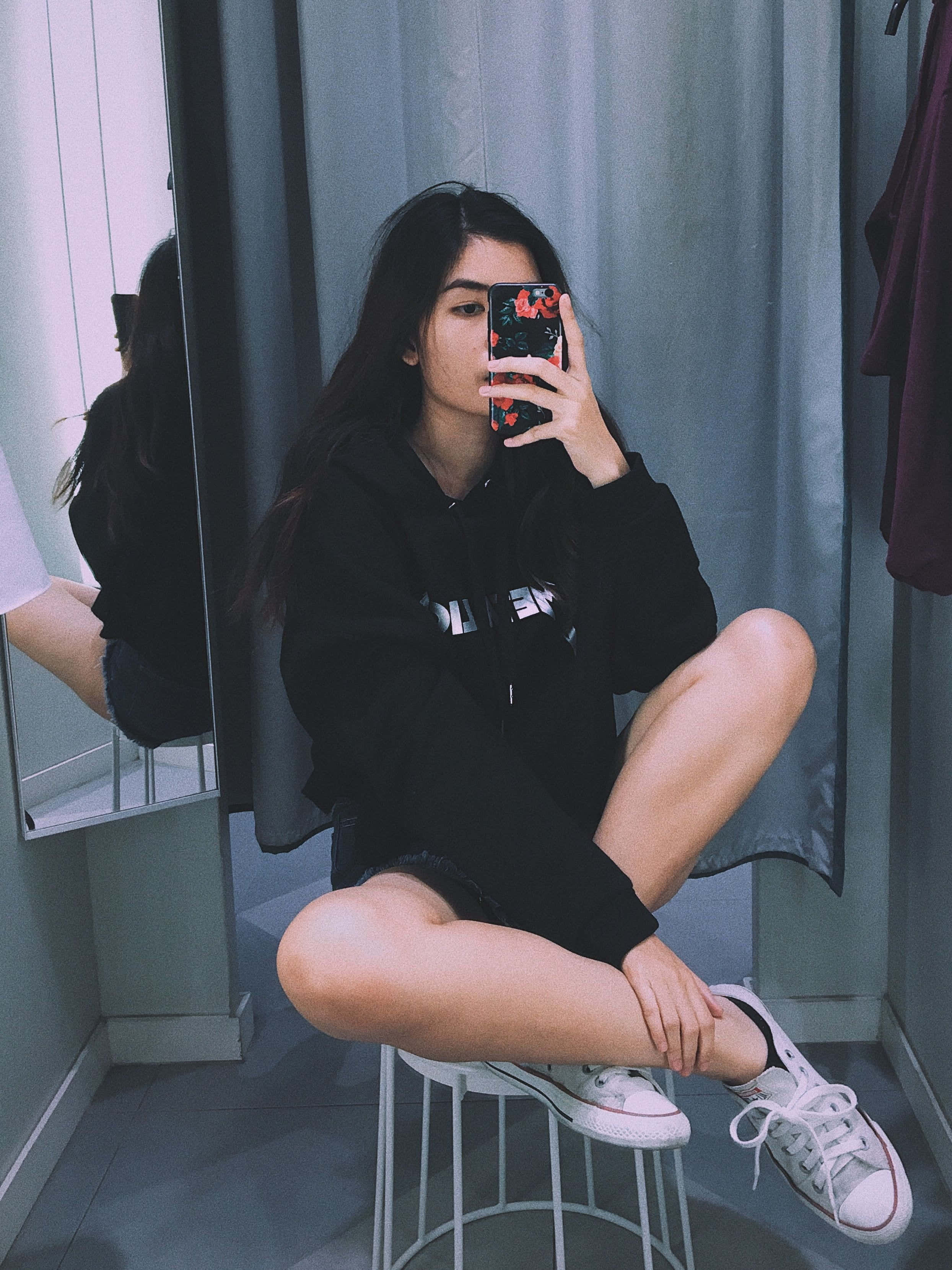 selfie 桌布 壁紙 — 一位年輕女性身穿黑色連帽衫和白色運動鞋，盤腿坐在凳子上，在試衣間對著鏡子自拍。