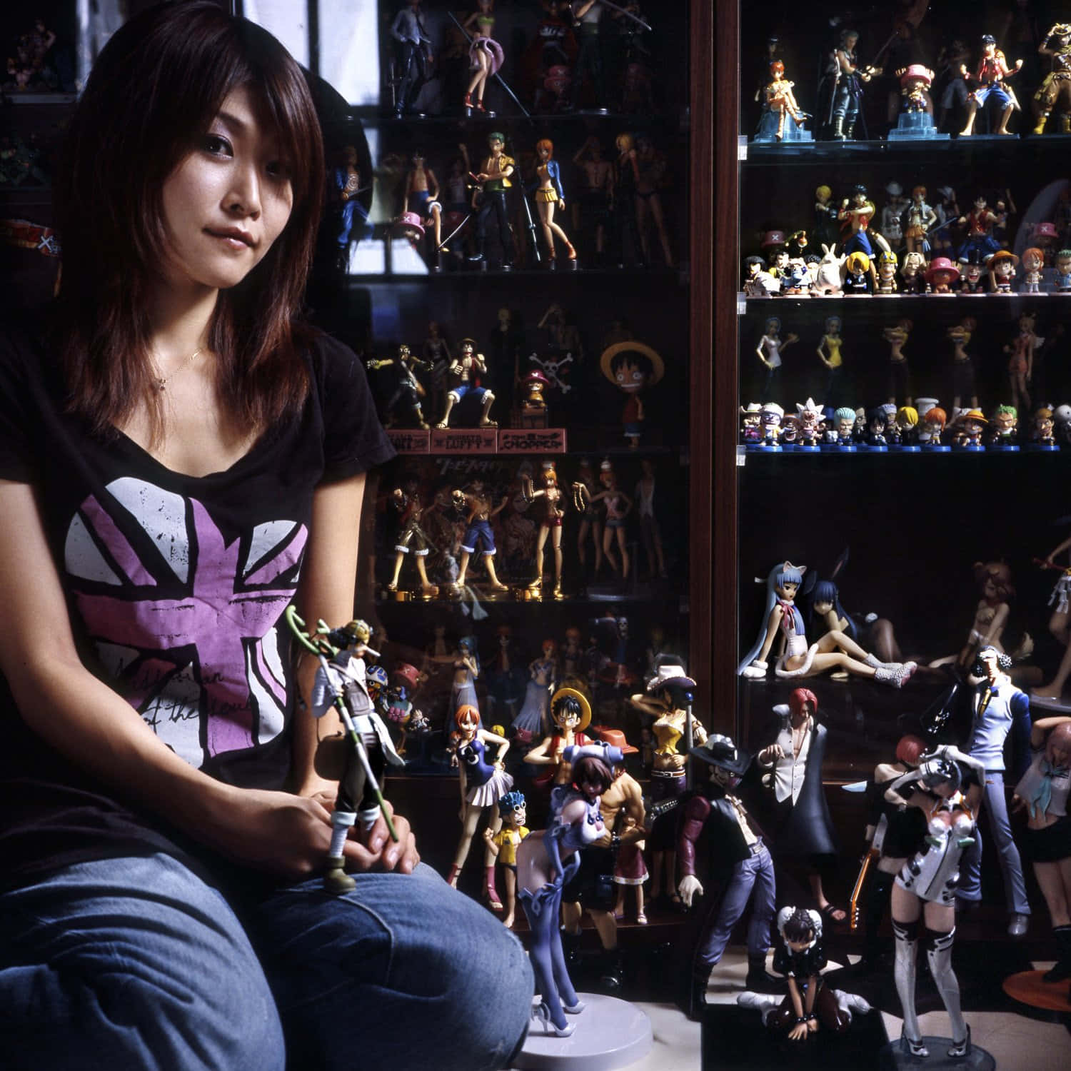 Otaku 桌布 壁紙 — 一位年輕女性靜靜地坐著，手持一個公仔，周圍是擺滿動漫角色收藏品的架子，房間燈光昏暗。