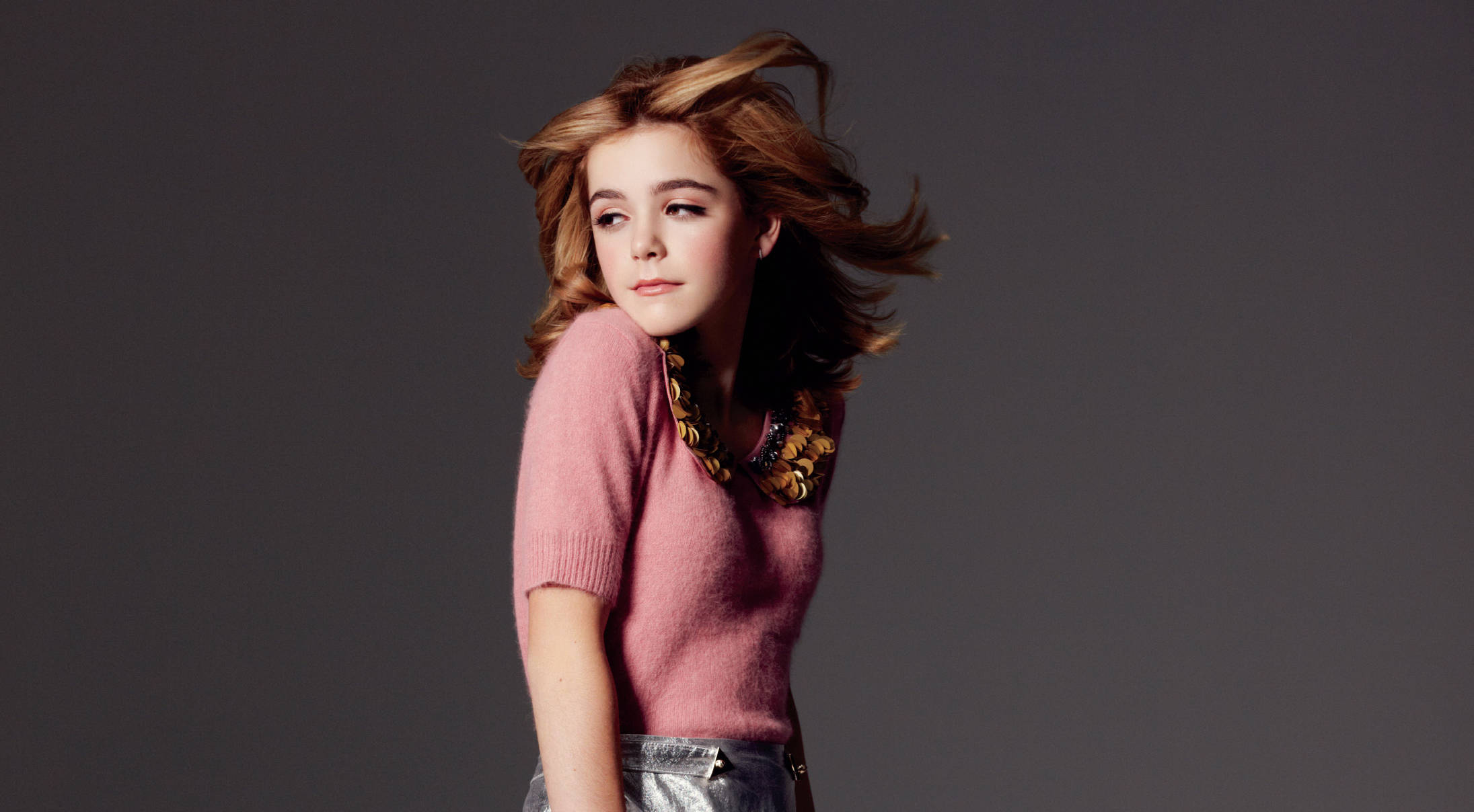 Kiernan Shipka 桌布 壁紙 — 一位年輕女性頭髮被風吹拂，身穿粉紅色毛衣和搶眼項鍊，背景為深灰色攝影棚。