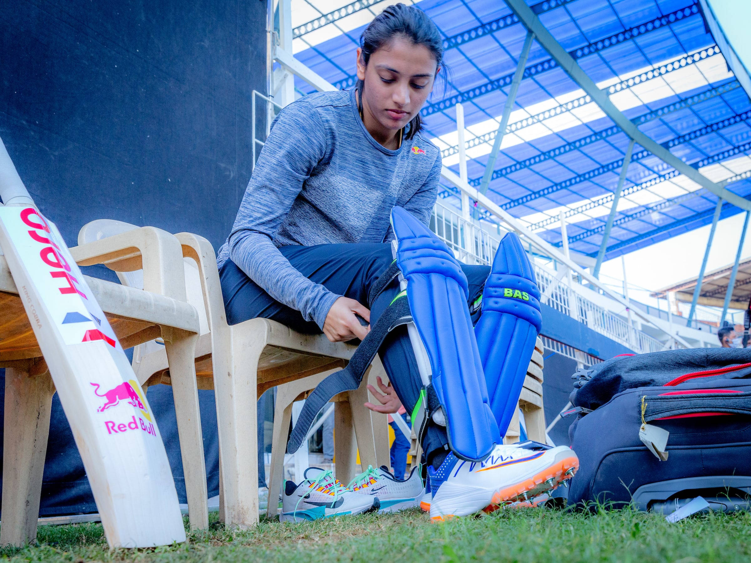 Smriti Mandhana 桌布 壁紙 — 一位年輕女板球手坐在長椅上，調整她的藍色護腿板，旁邊放著Red Bull板球拍和運動袋。
