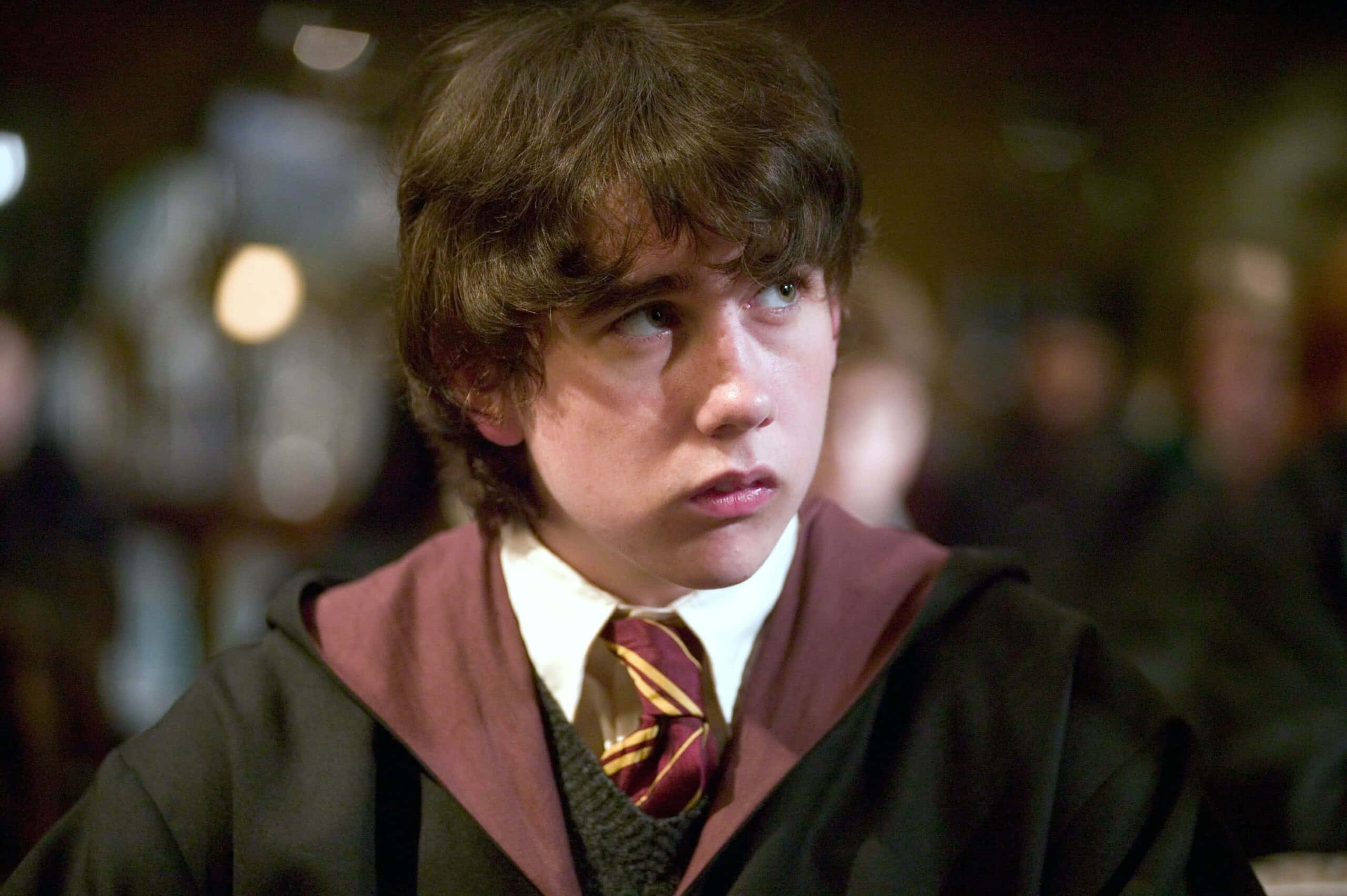 Neville Longbottom 桌布 壁紙 — 一位年輕巫師身穿栗色和金色長袍，在昏暗的魔法場景中帶著沉思的表情望向遠方。