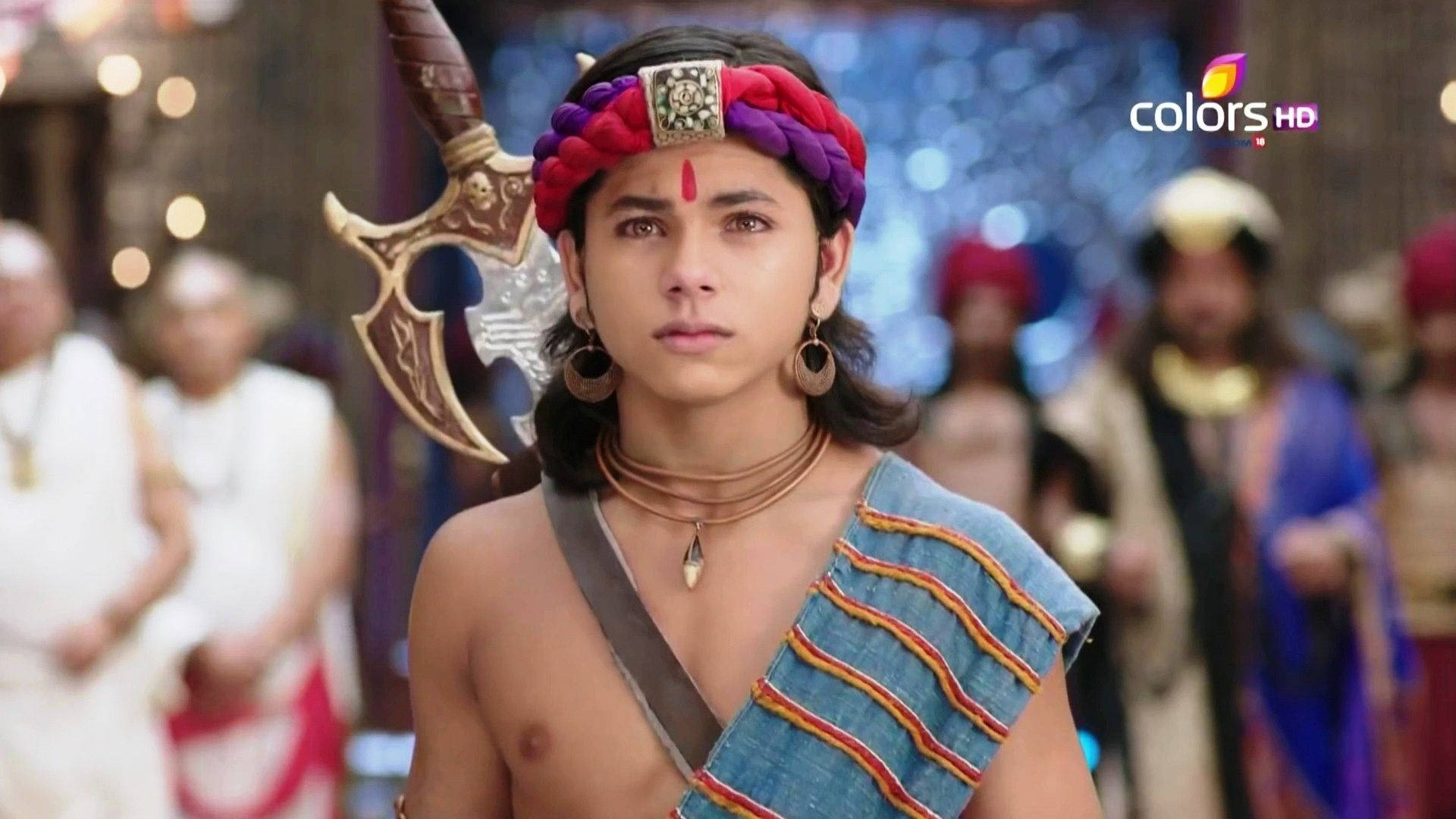 Siddharth Nigam 桌布 壁紙 — 一位年輕戰士戴著紅紫相間的頭帶，手持華麗的劍，站在一個有模糊侍從穿著傳統服飾的皇家宮廷中。