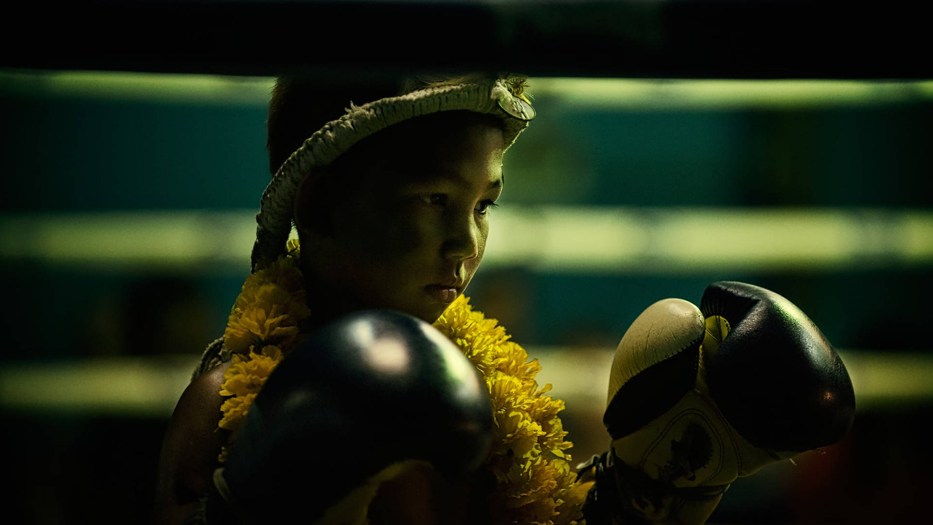 Boxing 桌布 壁紙 — 一位年輕拳擊手在燈光昏暗的擂台上，戴著黃色花環和黑色手套，專注地凝視前方。