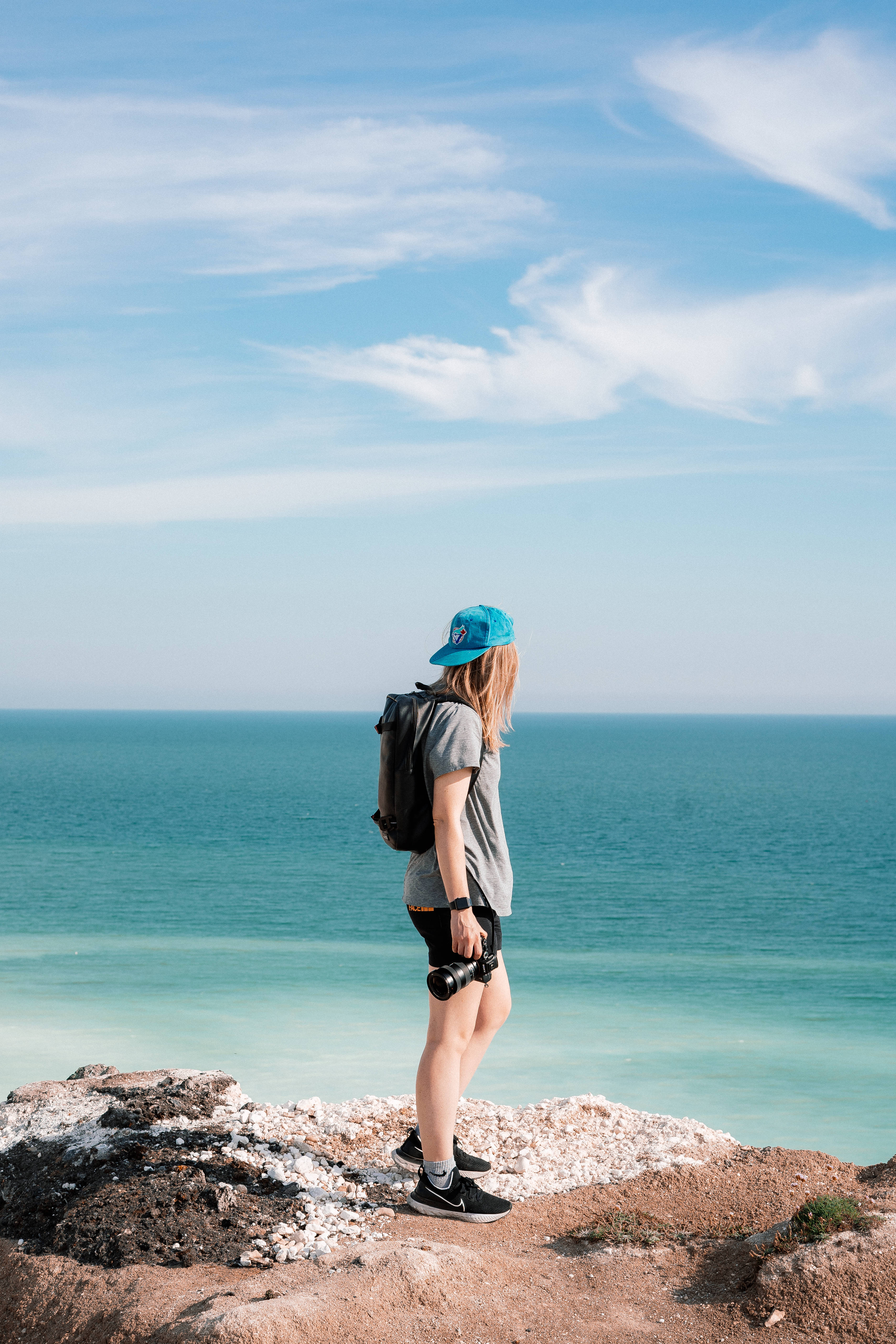 Tomboy 桌布 壁紙 — 一位年輕旅人背著背包和相機，站在岩石懸崖上，俯瞰著廣闊的碧綠海洋，天空湛藍，雲朵輕柔。