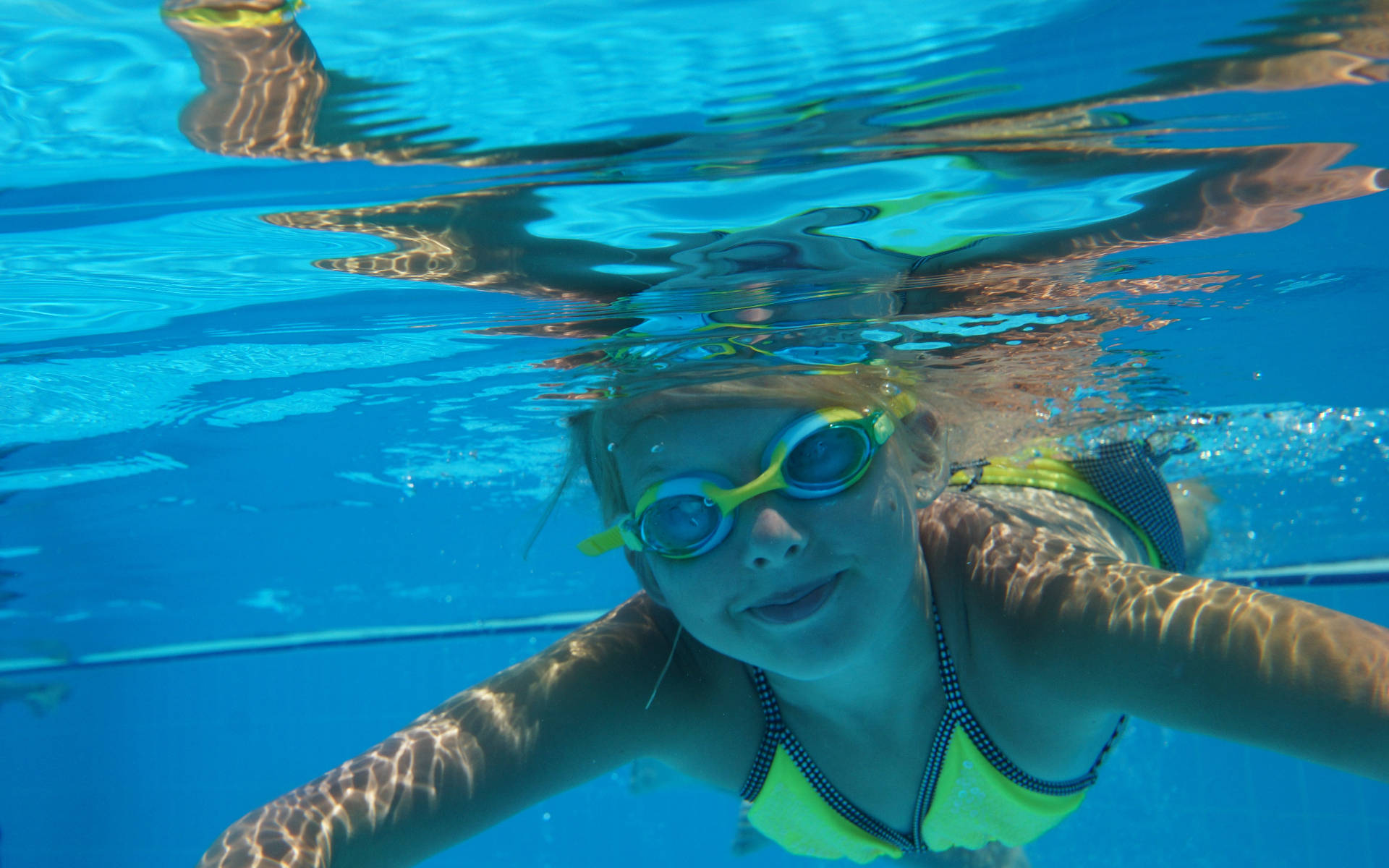 Swimming 桌布 壁紙 — 一位年輕泳者戴著亮黃色泳鏡和泳衣，在水下微笑，陽光在清澈的藍色泳池中泛起漣漪。