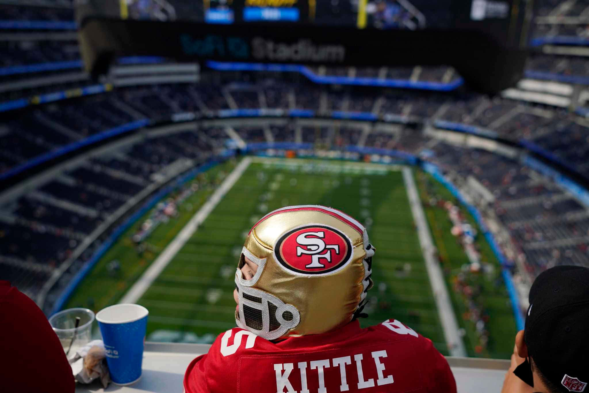 Sofi Stadium 桌布 壁紙 — 一位年輕球迷戴著金色頭盔和紅色球衣，在Levi's Stadium的上層看台觀看足球比賽。