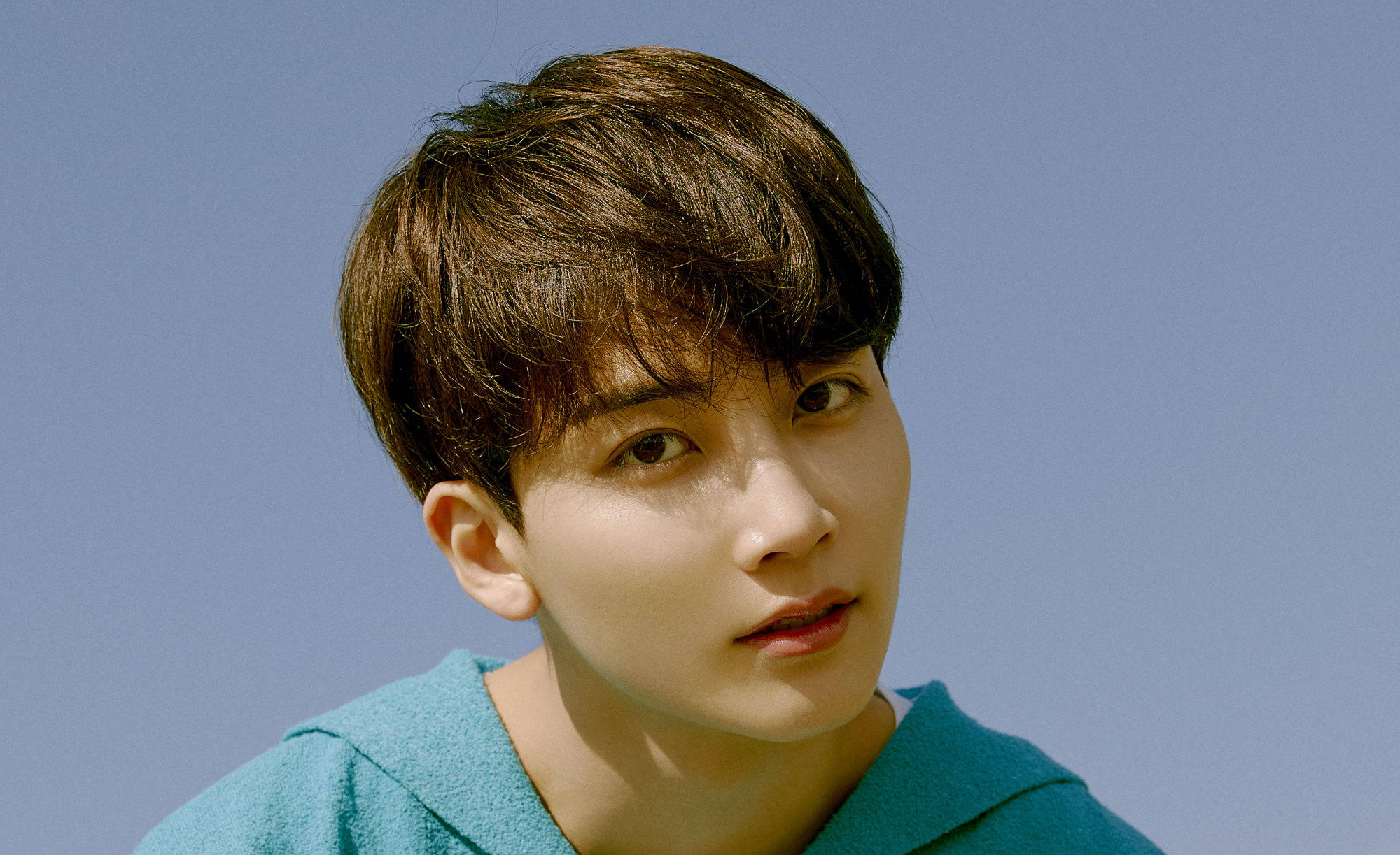 yoon jeonghan 桌布 壁紙 — 一位年輕男子，一頭凌亂的棕色頭髮，穿著青綠色連帽衫，在清澈的藍天下若有所思地凝視著。