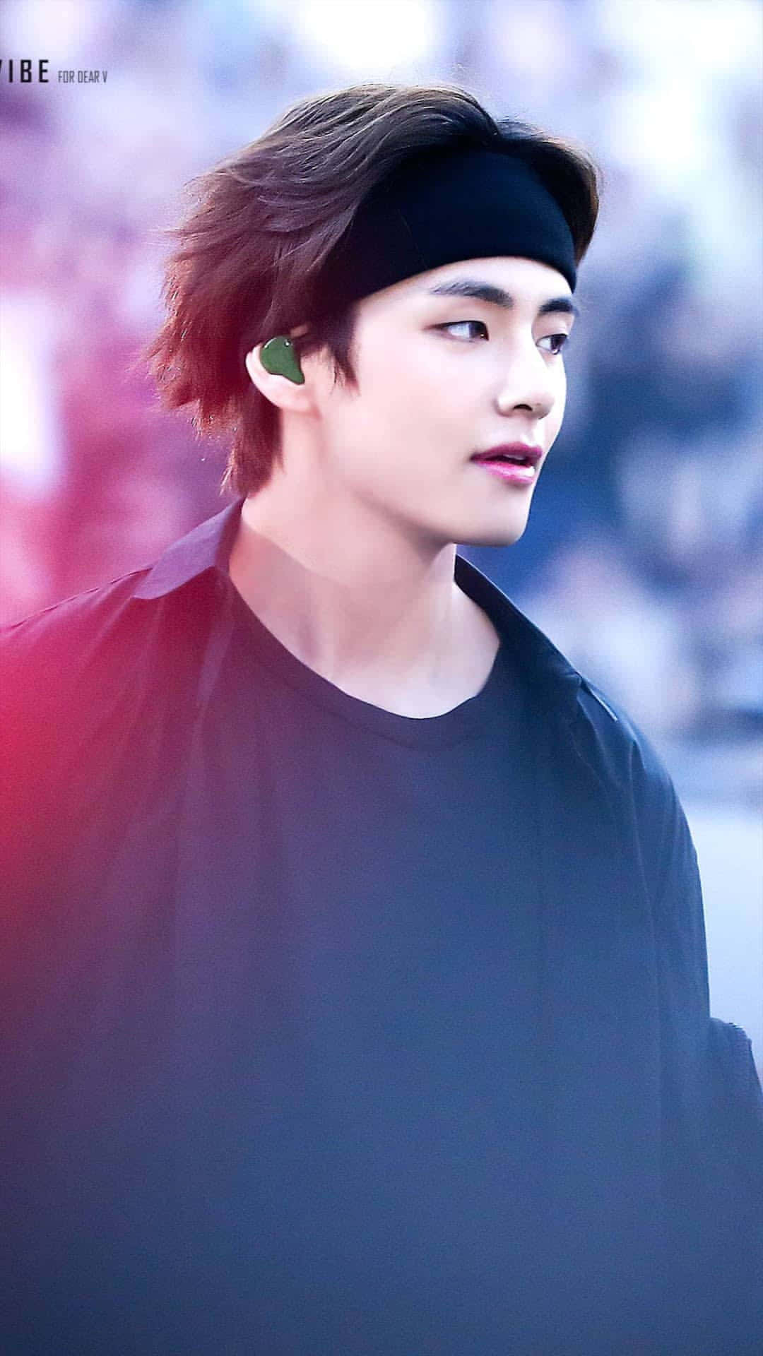 BTS 桌布 壁紙 — 一位年輕男子，一頭凌亂的棕色頭髮，戴著黑色頭帶，若有所思地望向一旁，身穿黑色襯衫，戴著綠色耳機，背景是柔和模糊的彩色。