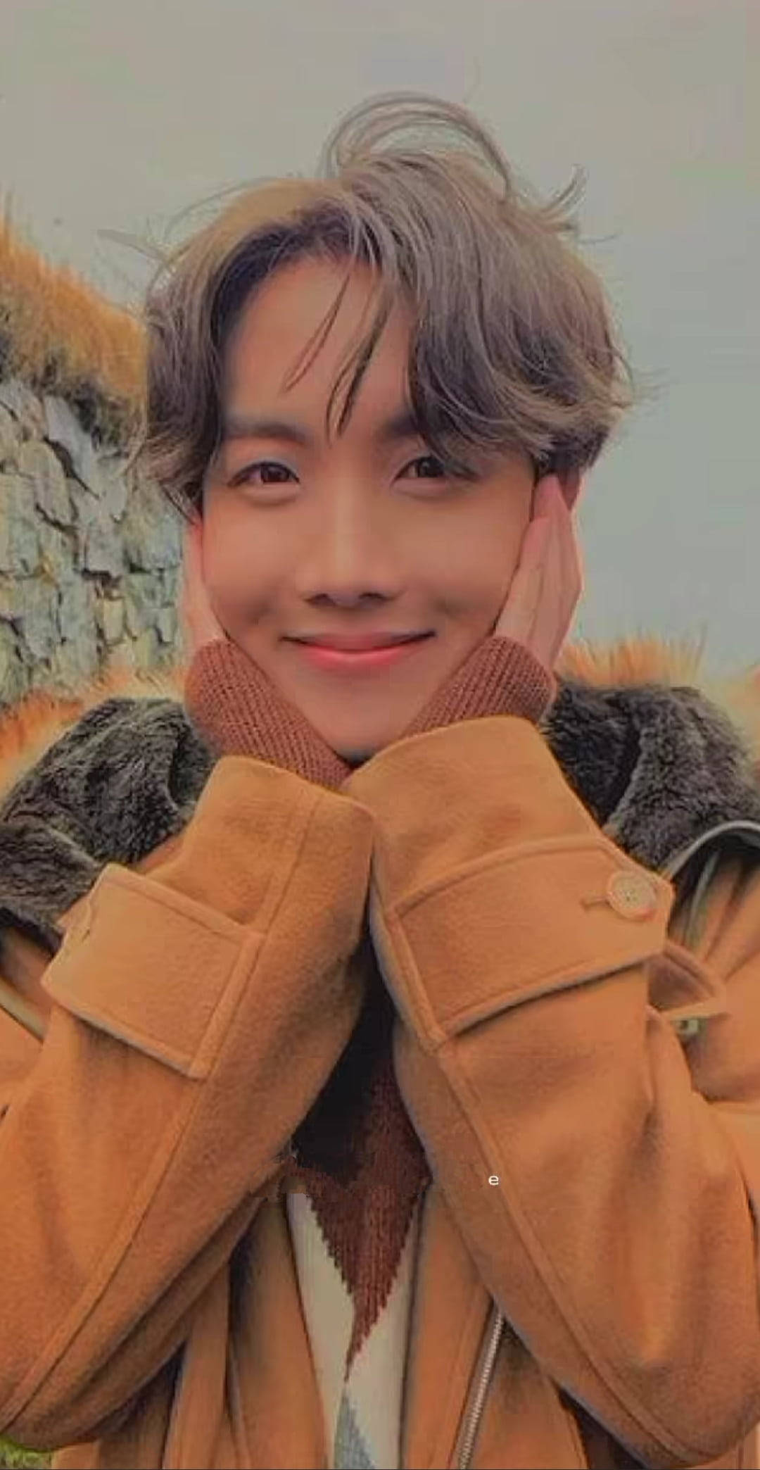 jhope 桌布 壁紙 — 一位年輕男子，一頭凌亂的灰髮，穿著帶毛領帽的棕褐色外套，在石牆背景下溫暖地微笑。