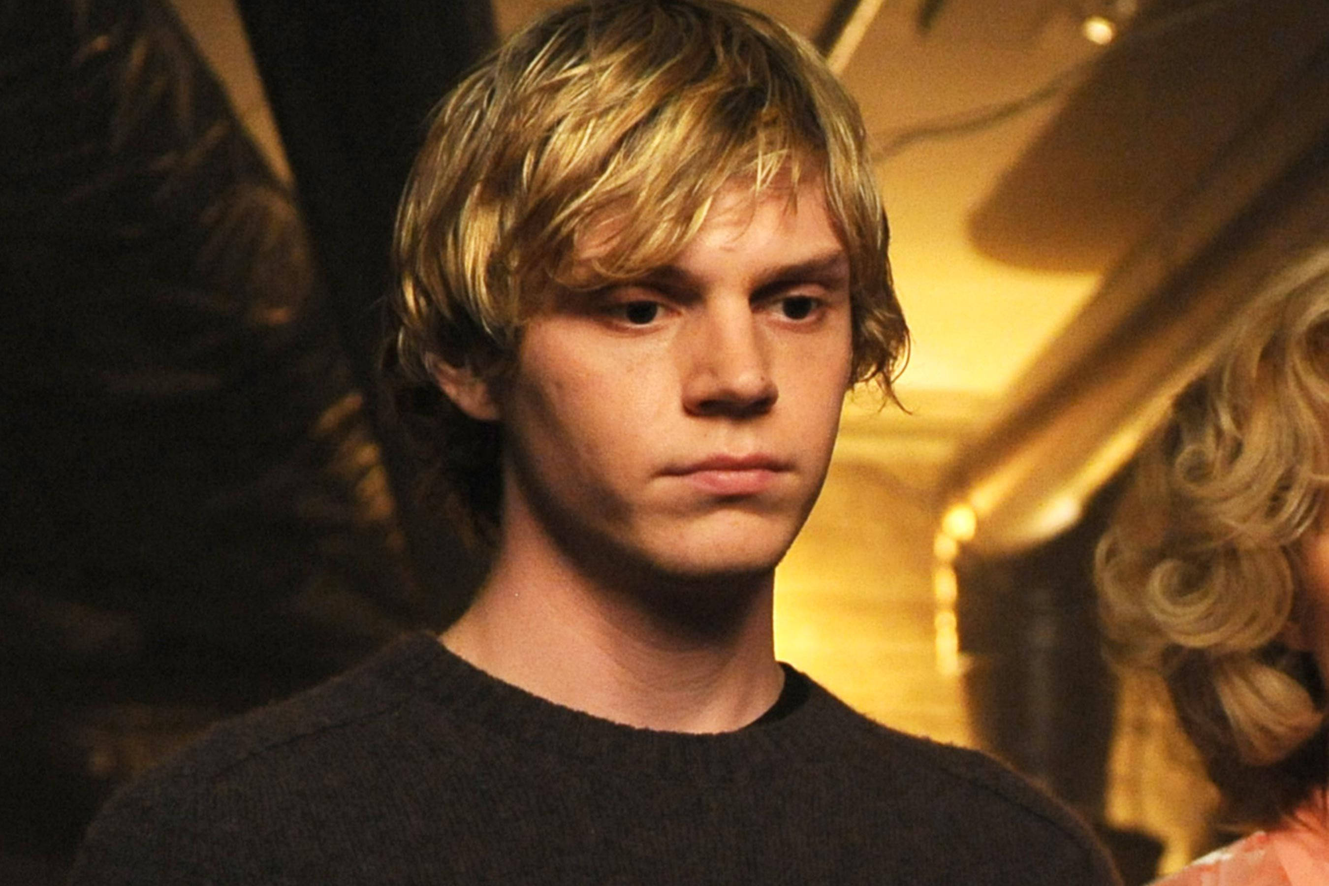 Tate Langdon 桌布 壁紙 — 一位年輕男子，一頭凌亂的金髮，在溫暖燈光下、柔和金色色調的室內環境中凝視著。