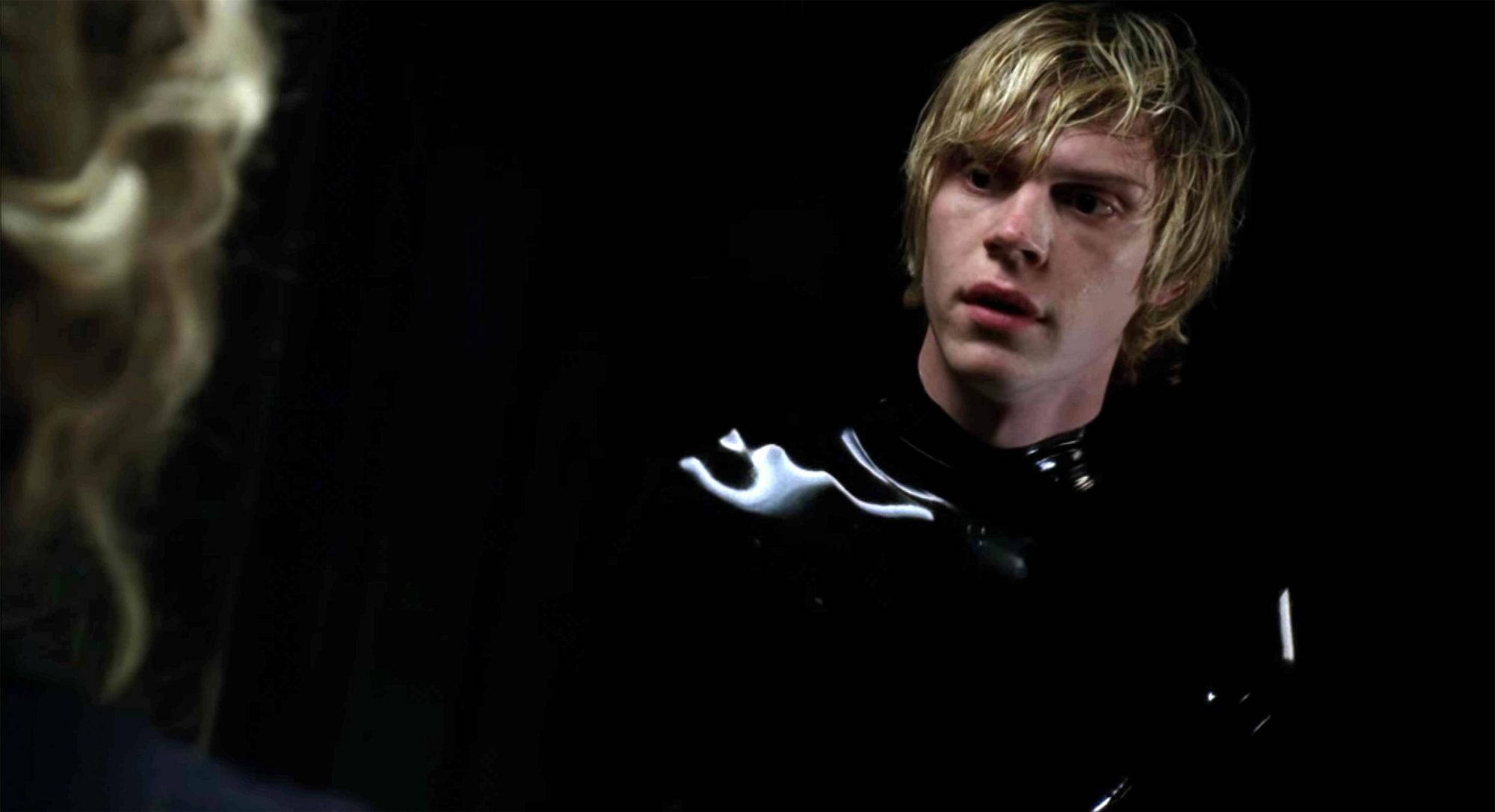 Tate Langdon 桌布 壁紙 — 一位年輕男子，一頭凌亂的金髮，在昏暗的場景中專注地凝視，身穿帶有白色發光裝飾的亮黑色服裝。